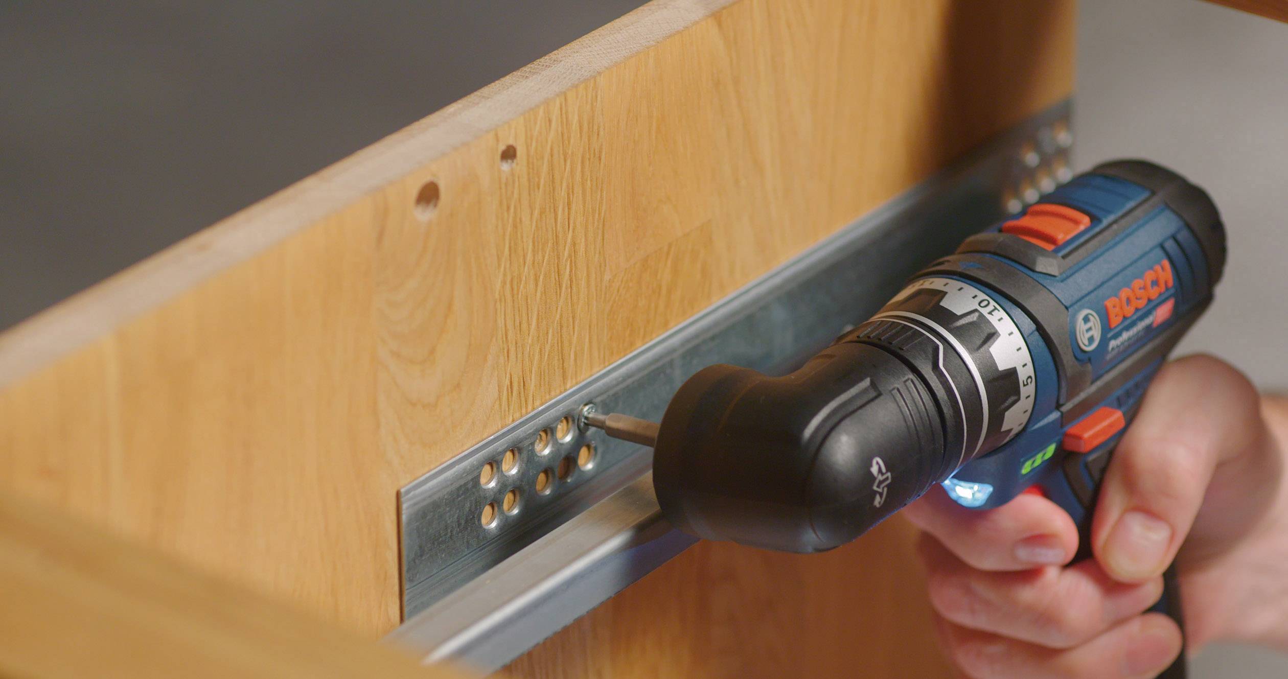 En hånd holder en blå akku-skruemaskine og fastgør en metalskinner på et stykke træ ved hjælp af skruer.