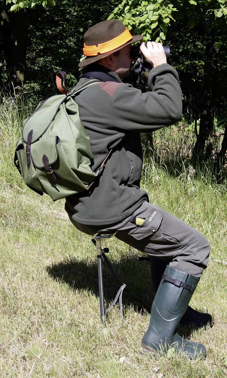 En person sidder på en skammel i det fri, iført jagtbeklædning og kigger gennem en kikkert. Personen har en stor grøn rygsæk.