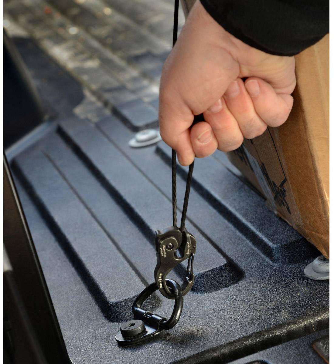En person holder en gummistrop med en krog, som er fastgjort til en sort overflade, sandsynligvis til at sikre et objekt.