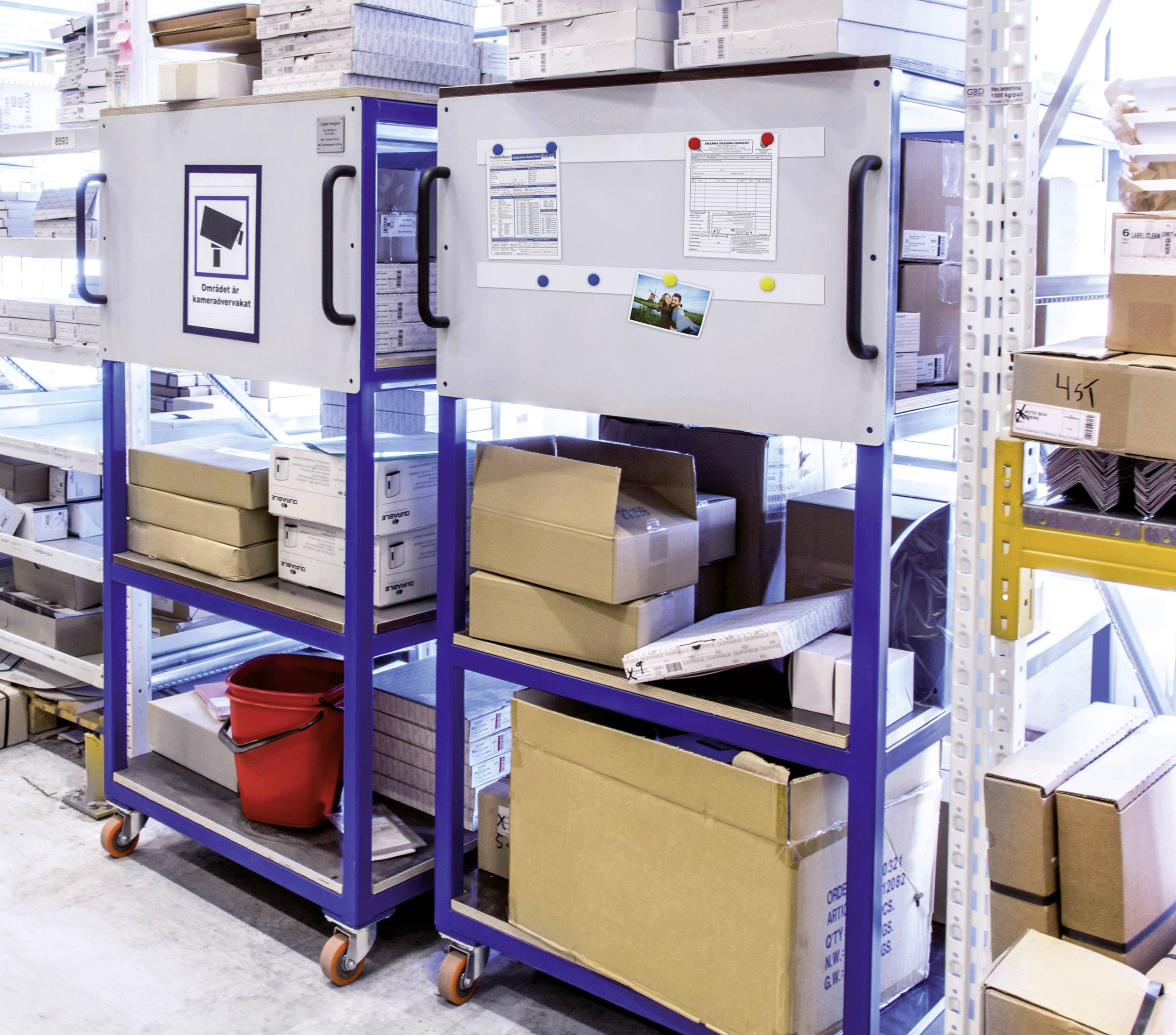 To metalblå reoler på hjul i et lager. De er fyldt med pakkasser, en rød spand, papirer og opslagstavler.