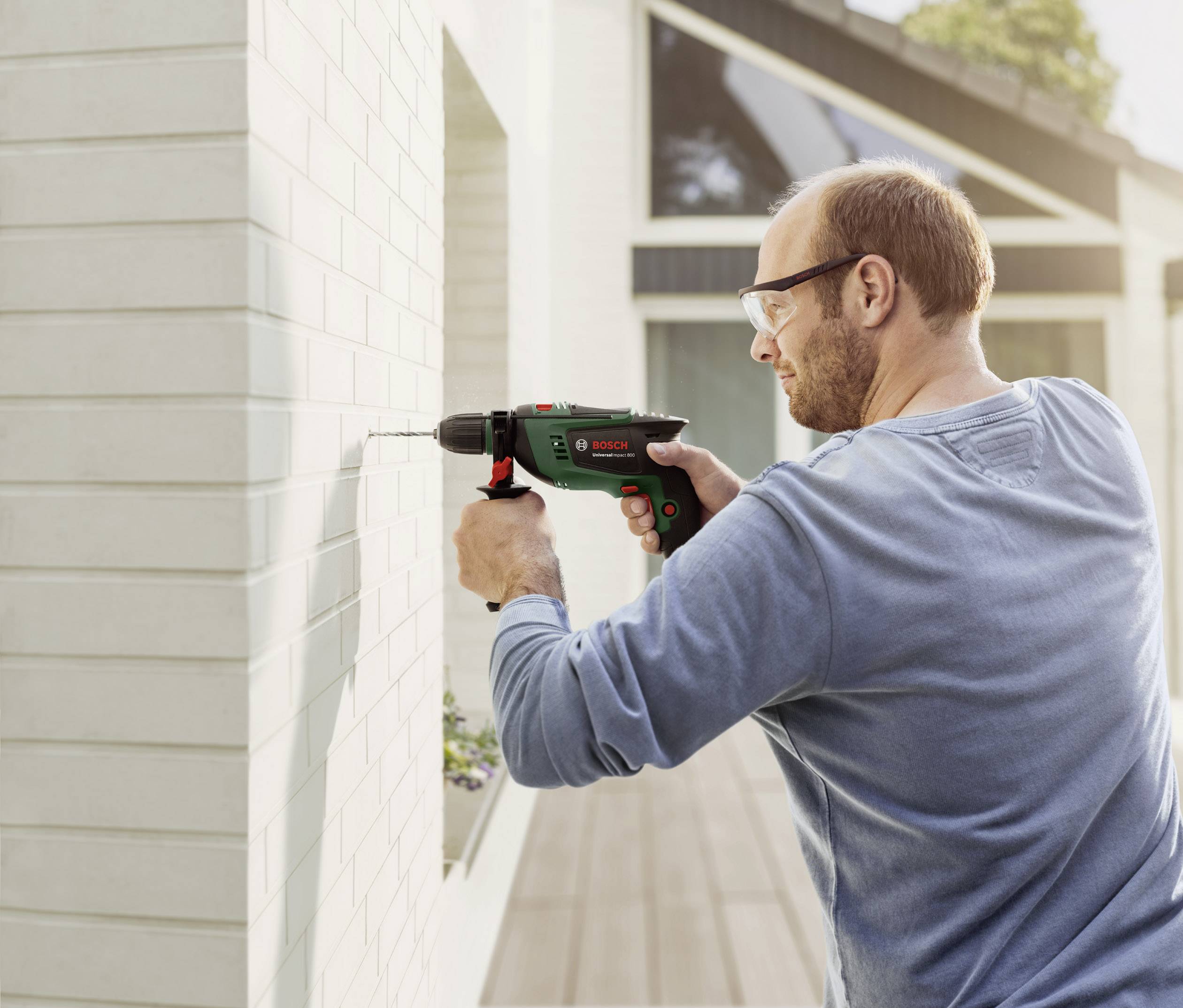 En mand borer med en grøn boremaskine i en hvid flisebelagt væg. Han bærer sikkerhedsbriller og står på en terrasse.