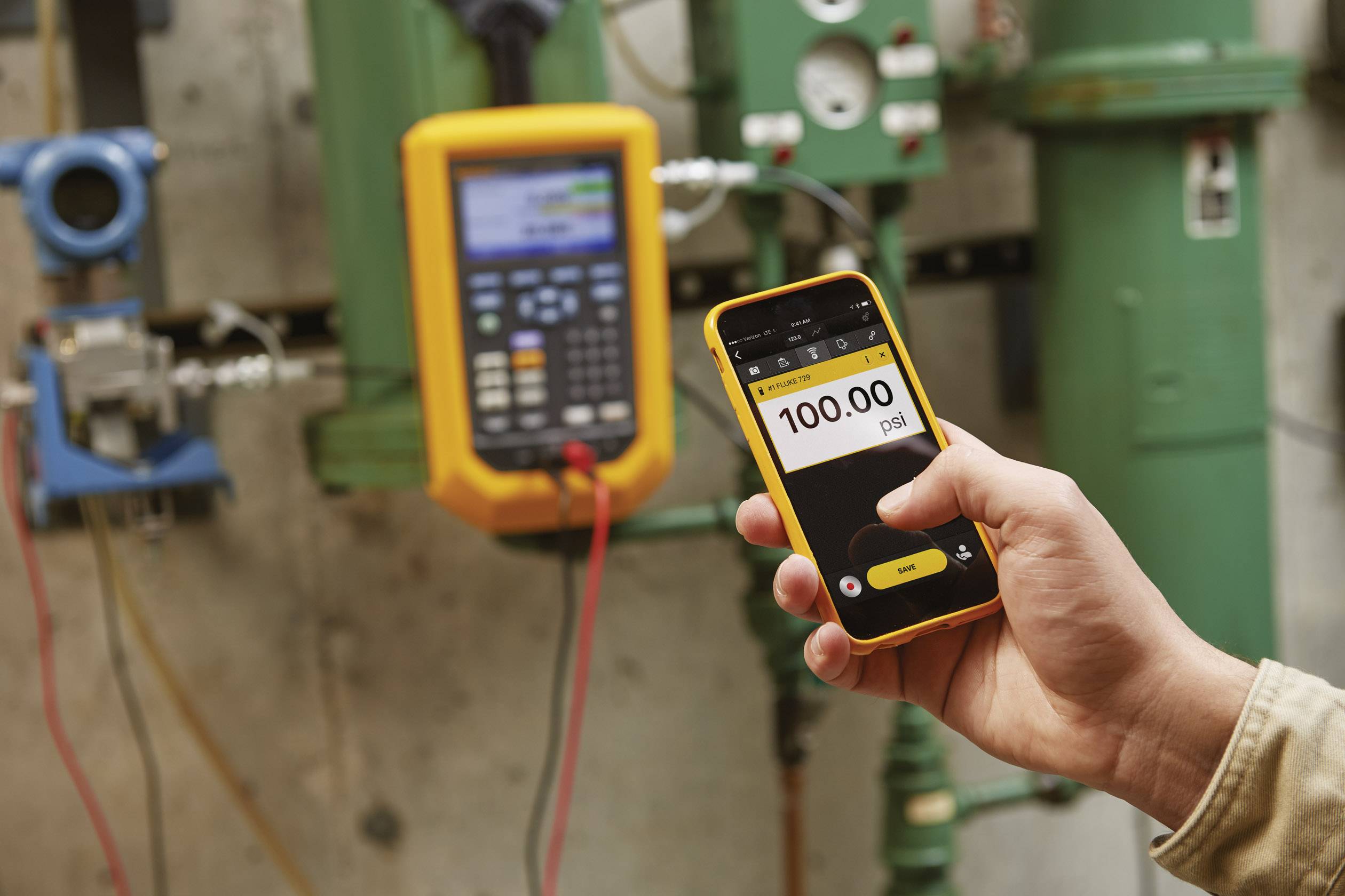 En hånd holder en smartphone, som viser et trykdisplay på '100,00 psi'. I baggrunden ses tekniske apparater.