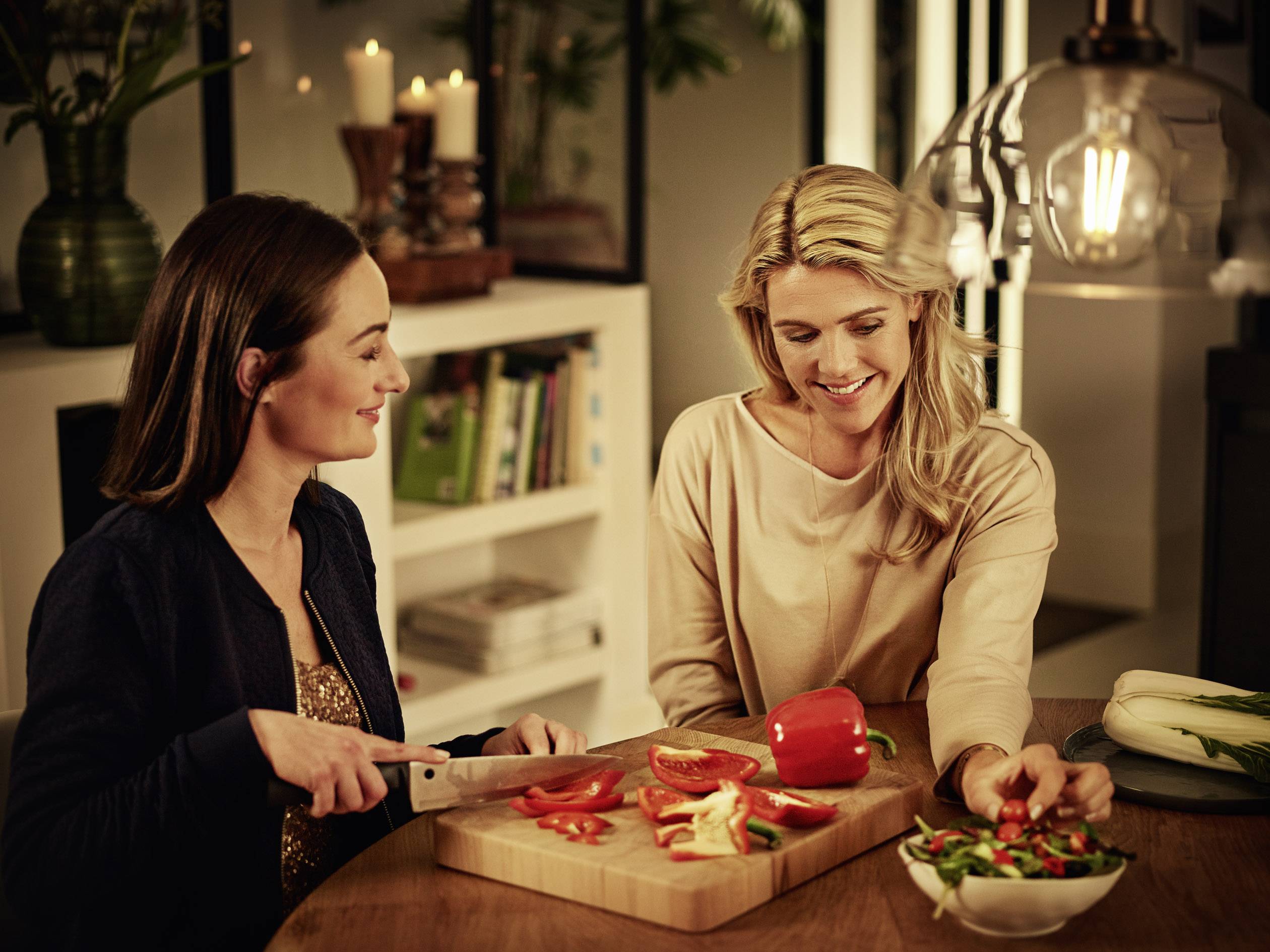 To kvinder forbereder peberfrugt og salat i et hyggelig køkken, smiler og taler sammen. En af dem skærer peberfrugt på et træskærebræt.