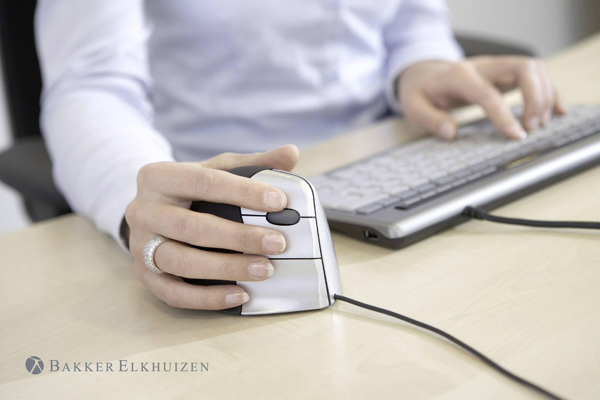 En person holder en ergonomisk mus i højre hånd og taster på et tastatur. Ergonomisk design til behagelig brug.
