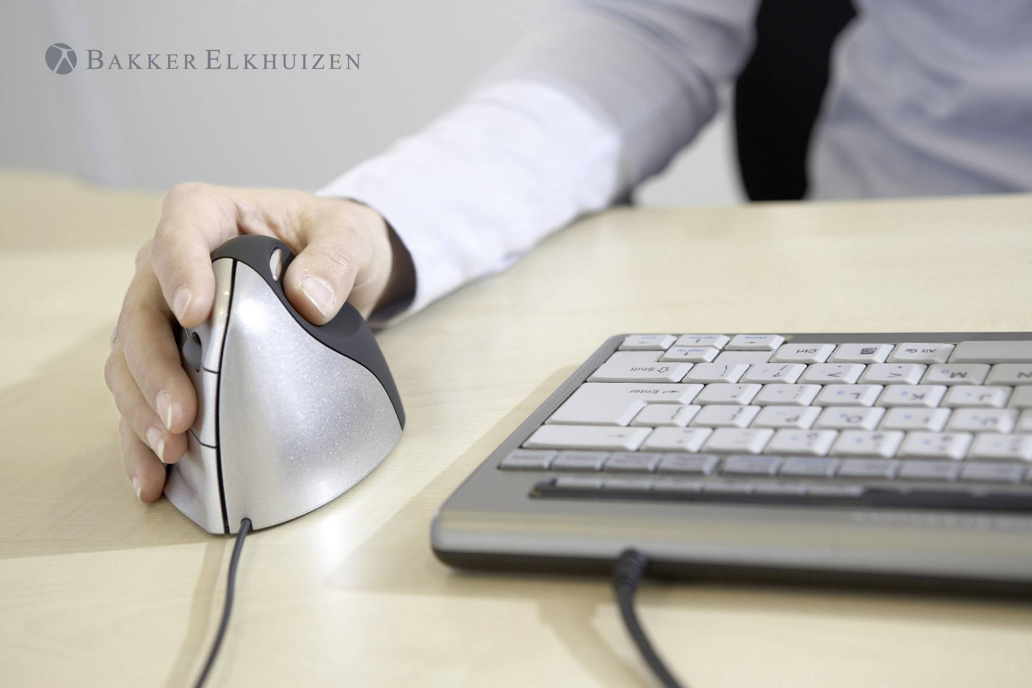 En person bruger en ergonomisk, vertikal computermuse ved siden af et tastatur på et bord.