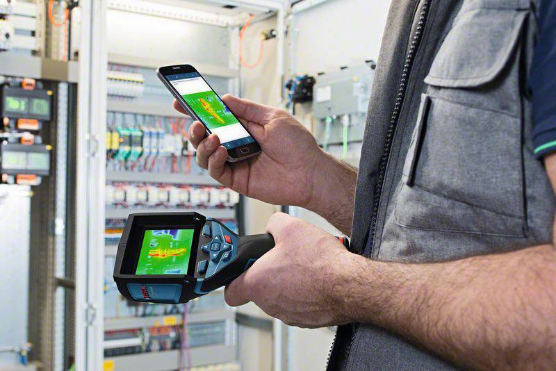En tekniker holder et digitalt diagnosticeringsapparat og en smartphone foran et åbent el-tavleskab og kontrollerer elektriske anlæg.