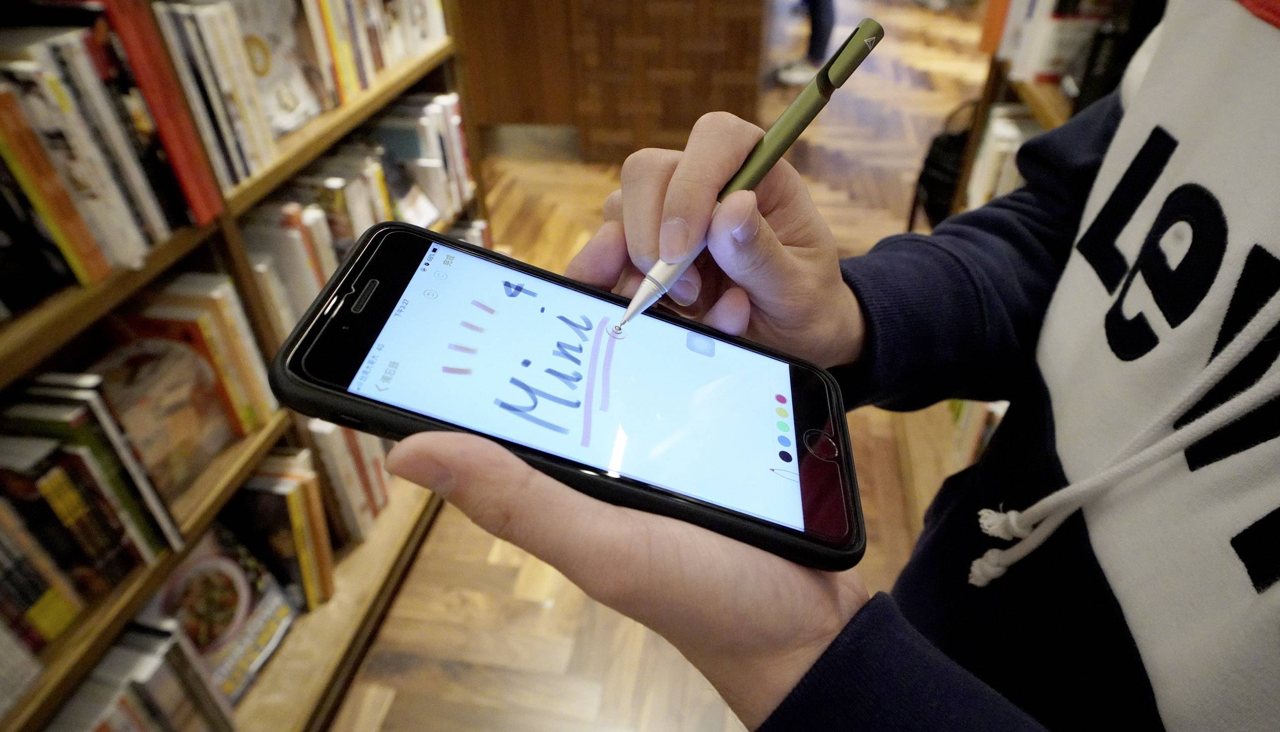 En person bruger en stylus til at skrive 'Mini' på en smartphone. I baggrunden er der bogstativer synlige.