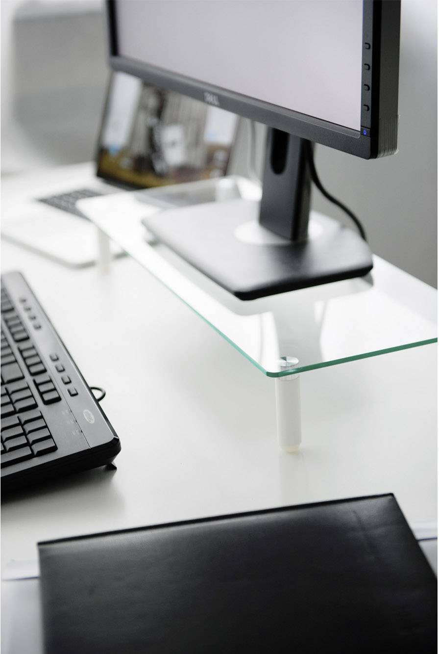 En computerskærm står på en glashylde på et skrivebord, ved siden af ligger et tastatur, og en bærbar computer er placeret i baggrunden.
