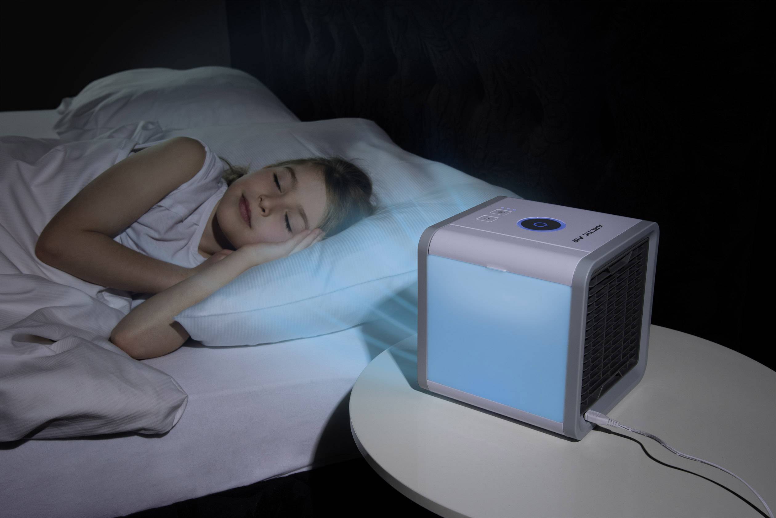 En person sover fredeligt i sengen, mens en blå bærbar aircondition står tændt på natbordet ved siden af.