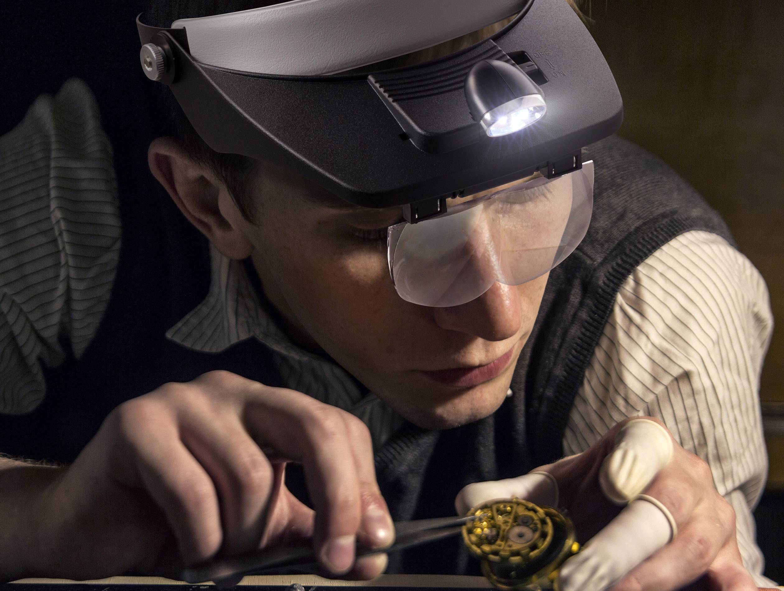 En person arbejder fokuseret på en lille, mekanisk genstand og bærer en forstørrelsebrille med integreret lys.