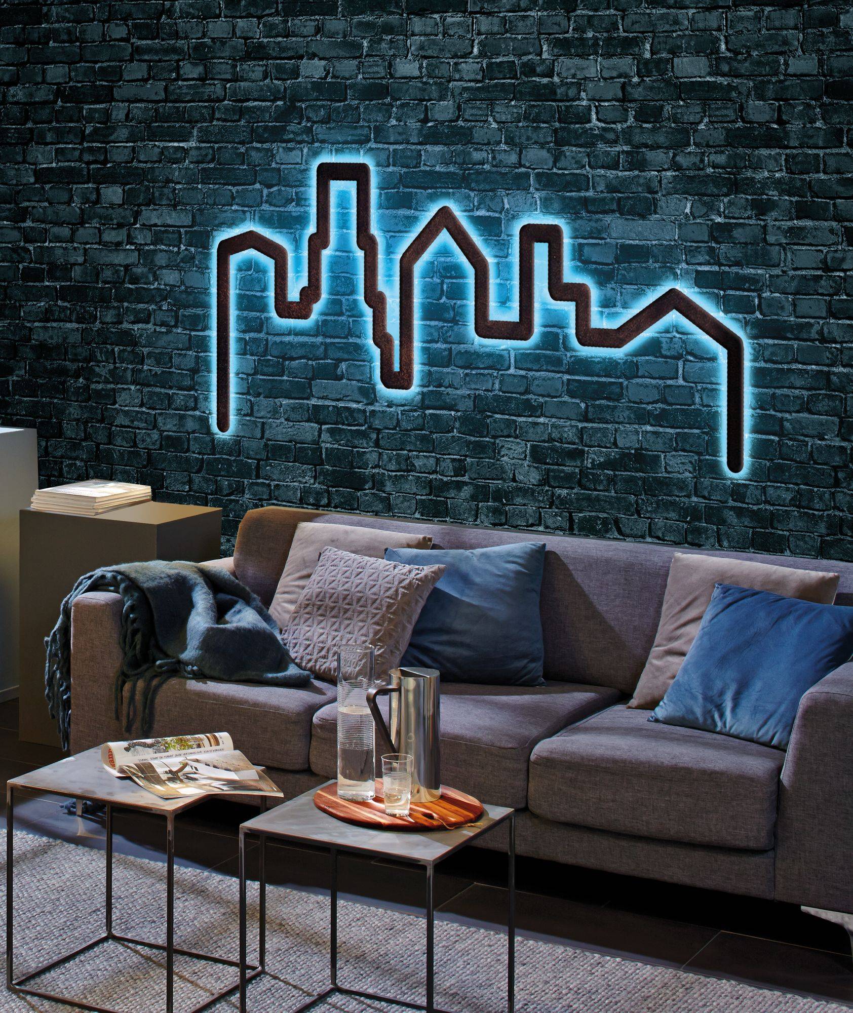 Et moderne stue med en grå sofa, blå puder og neon-vægkunst, der viser en bysilhuet på en murstensvæg.