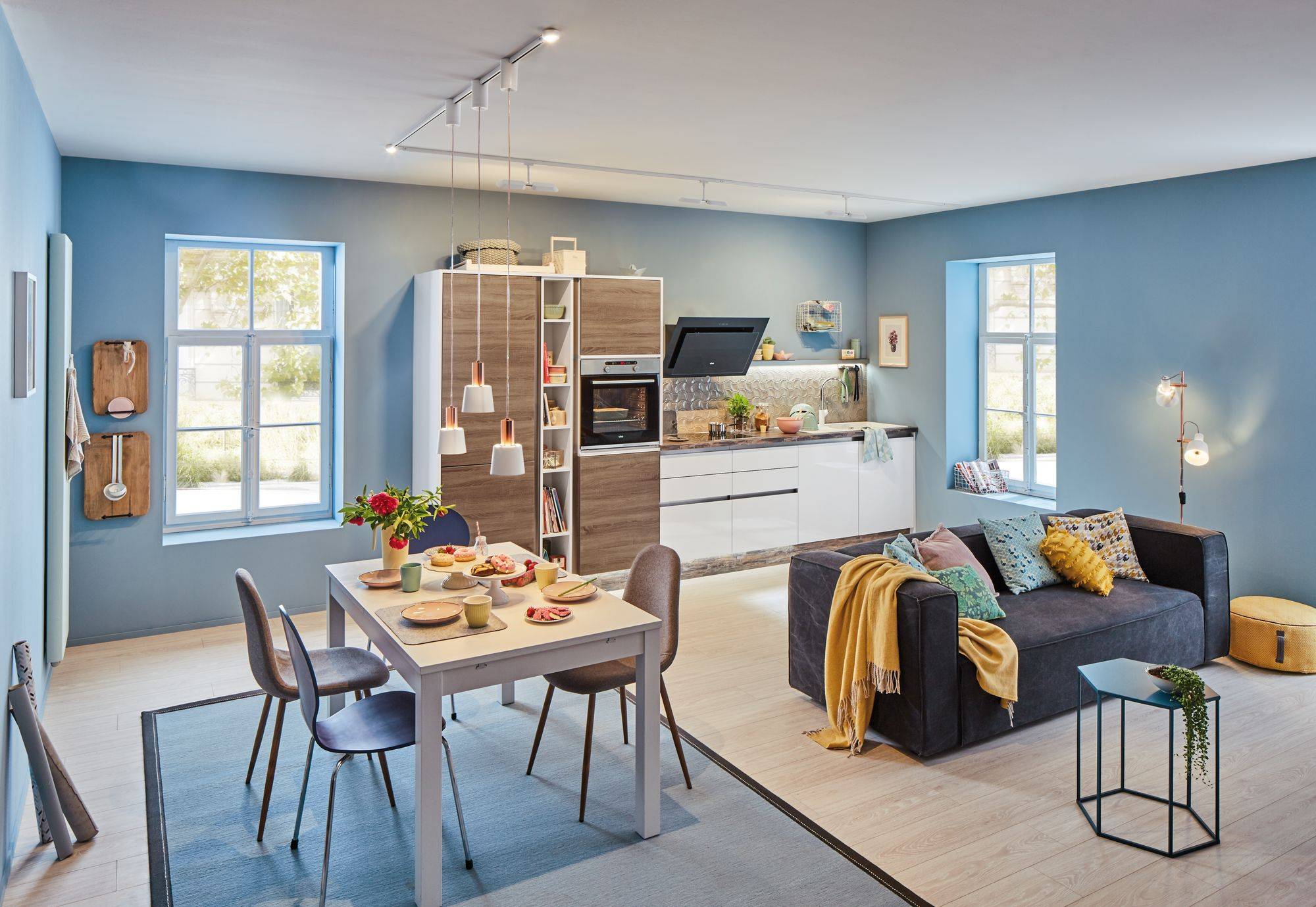 Et hyggeligt opholds- og køkkenrum i ét, med et spisebord, stole, sofa, et moderne køkken og pastelblå vægge, oplyst af dagslys.
