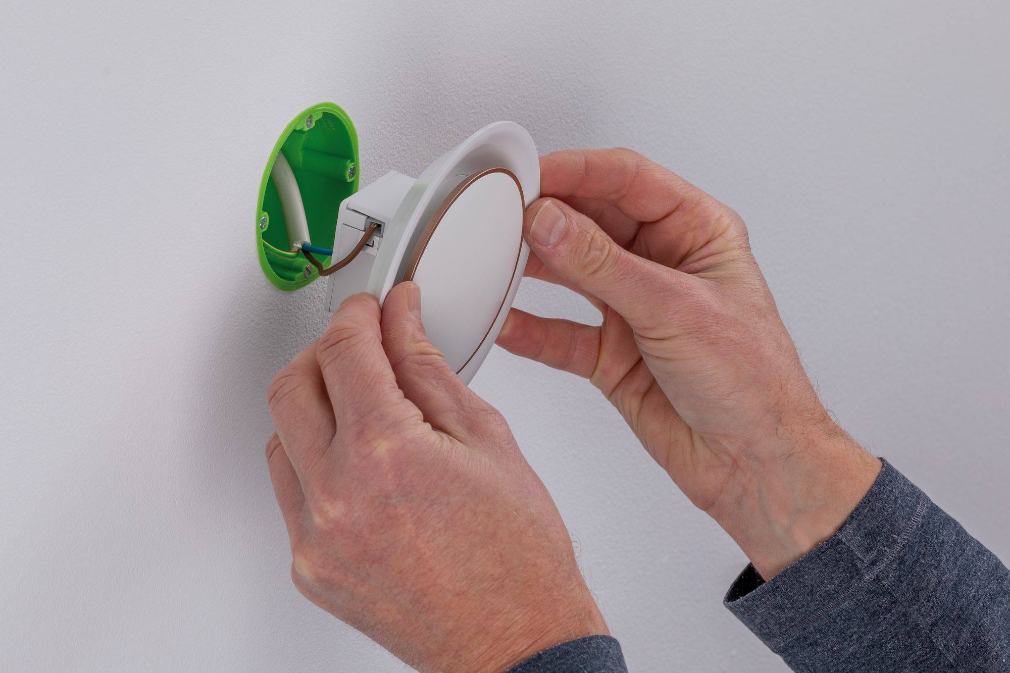 En person installerer en rund LED-lampe i en grøn, åben stikkontakt på en hvid væg.