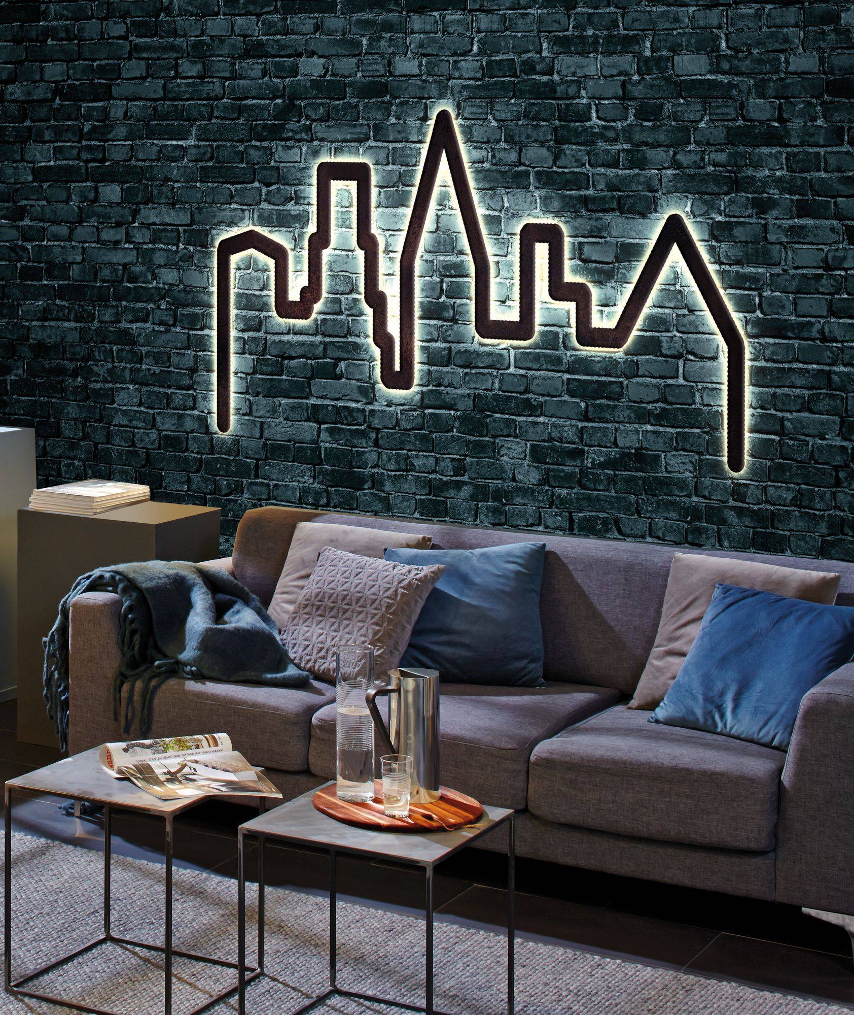 Moderne boligindretning med grå sofa, blå puder og tæppe. Væg med murværksudseende og oplyst skyline-kontur som dekoration.