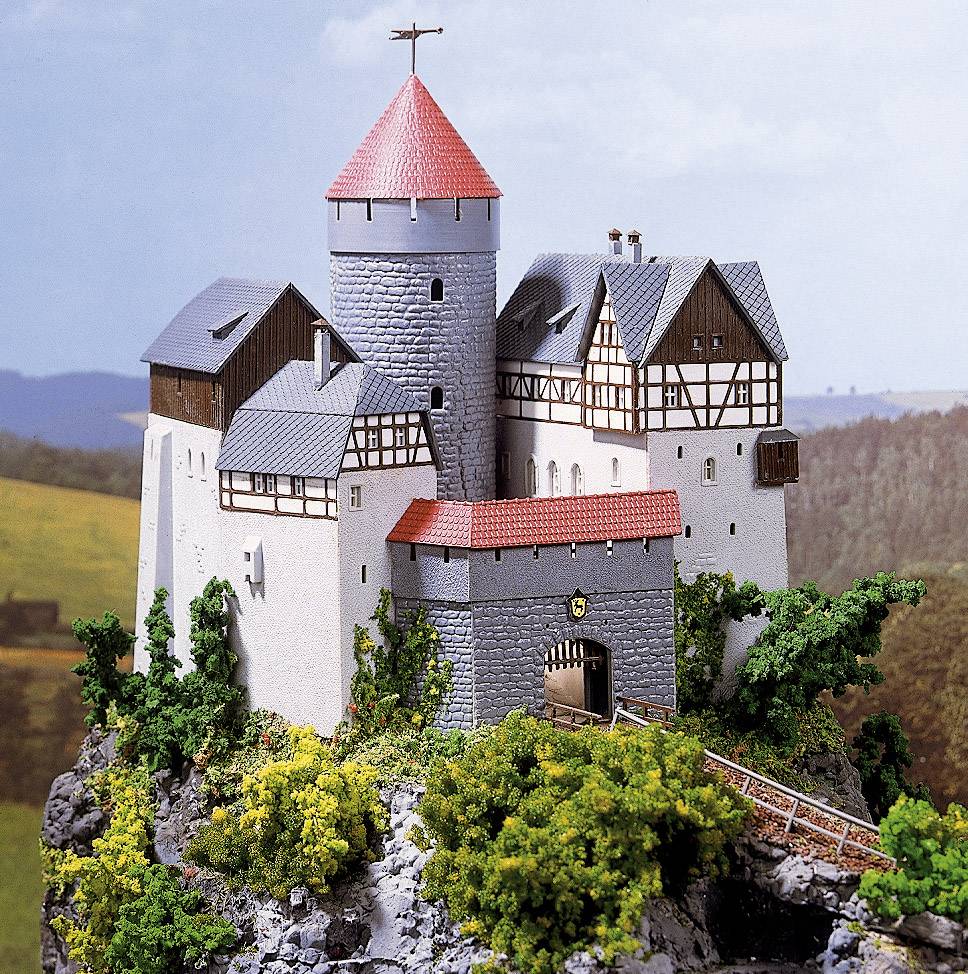 En detaljeret miniatureborg står på en klippefuld bakke, omgivet af træer. Borgen har et rundt tårn og røde tage.