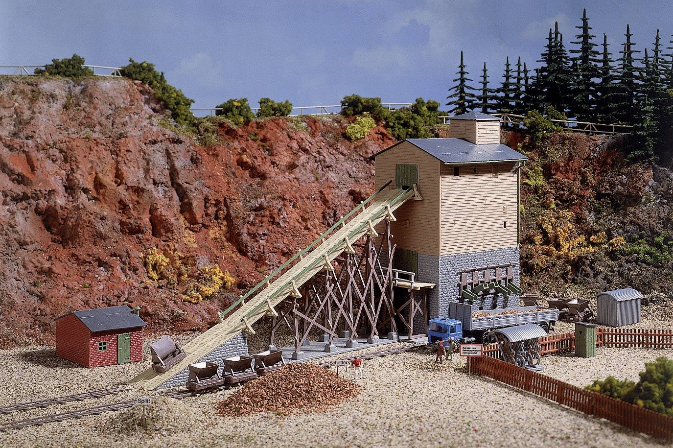 En miniature modelbanejlandskab med en brun bygning, der flankeres af en skrårampe med et transportbånd; omgivet af vegetation.