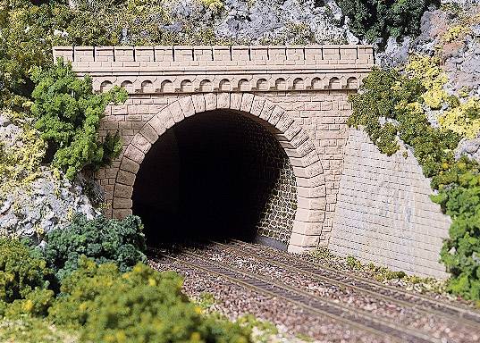 En modelbanetunnel lavet af mursten, omgivet af grøn vegetation; skinnerne fører ind i den mørke åbning.