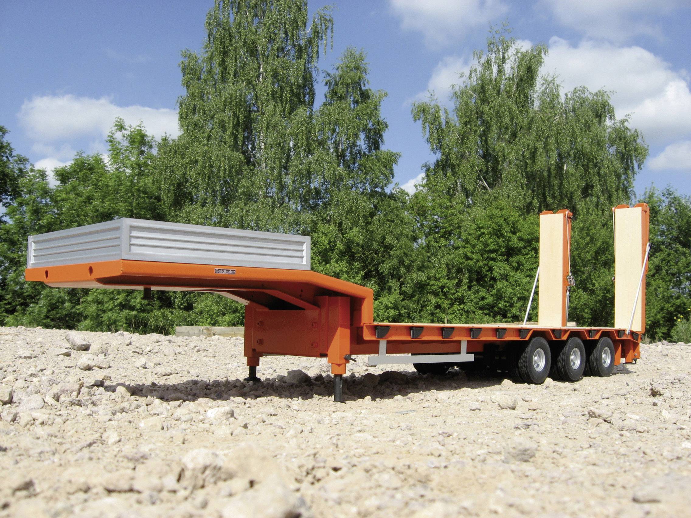 En orange sættevogn med ramper på en tør, sandet overflade. I baggrunden ses grønne træer.