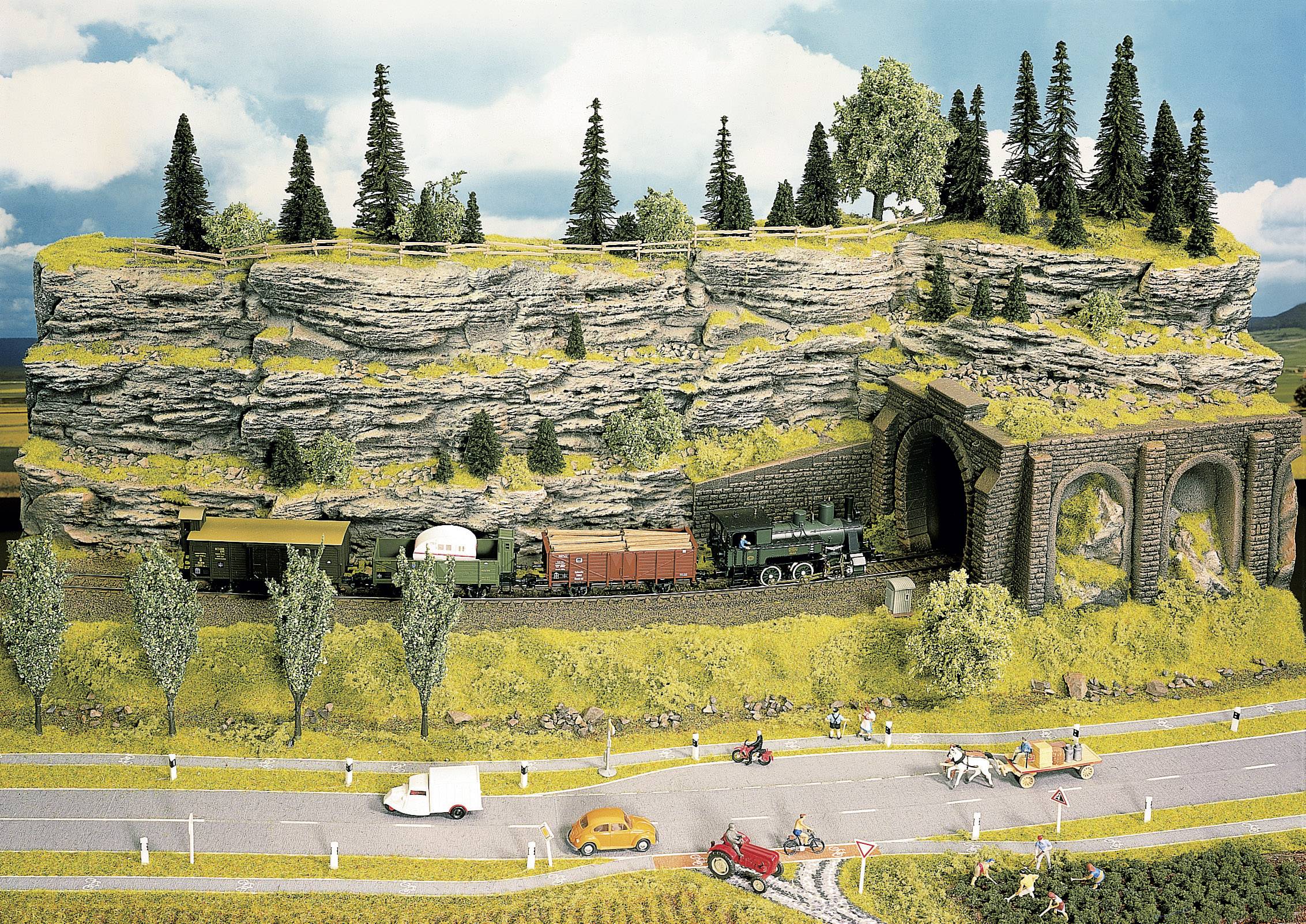 Modeltogslandskab med et tog, der kører gennem en tunnel i et bjerglandskab bevokset med skov. En vej ses i forgrunden.