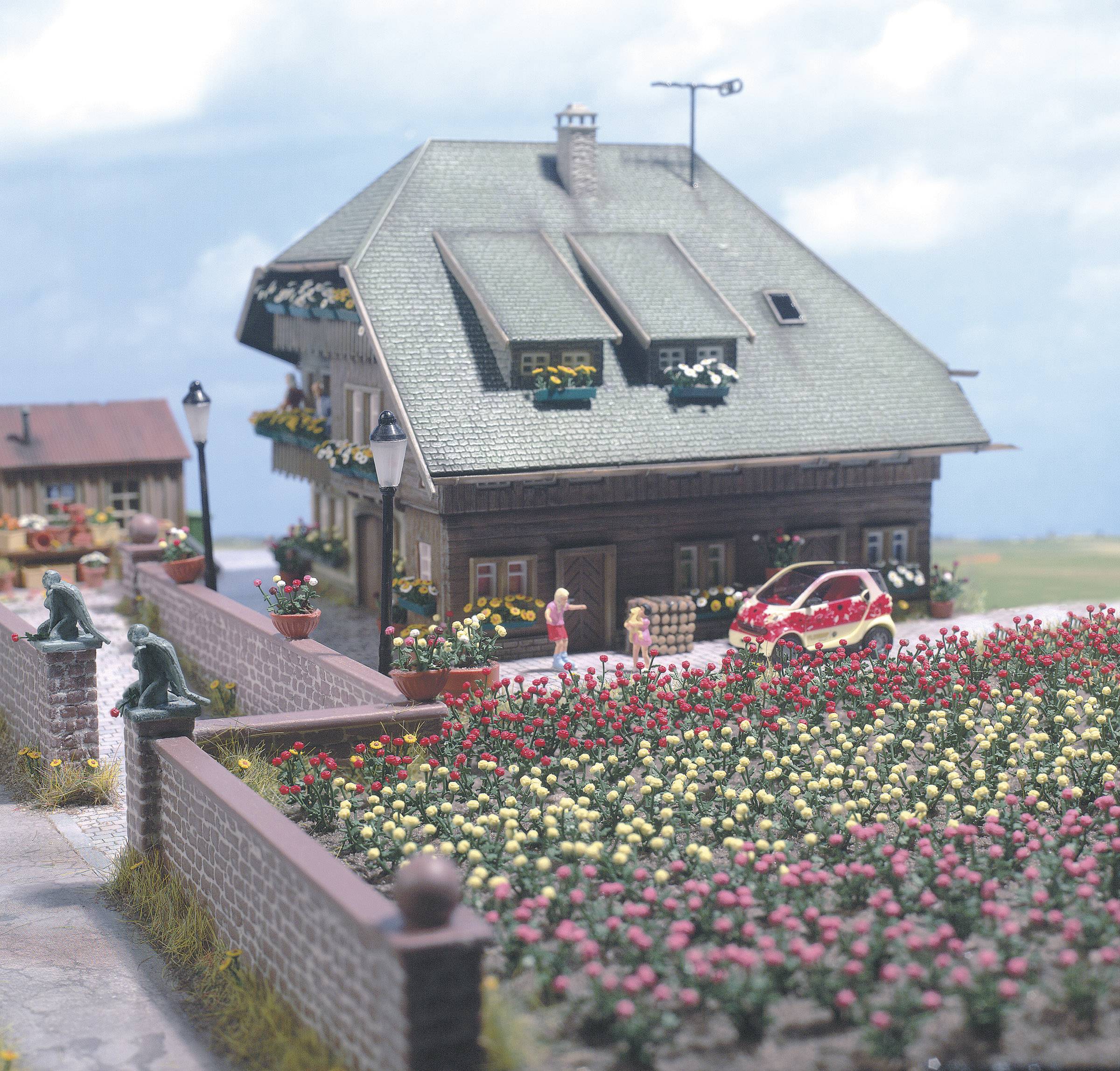 Landligt hus med sadeltag, blomsterbede i forgrunden og en rød figur, der står ved siden af en rød bil. Klar himmel i baggrunden.