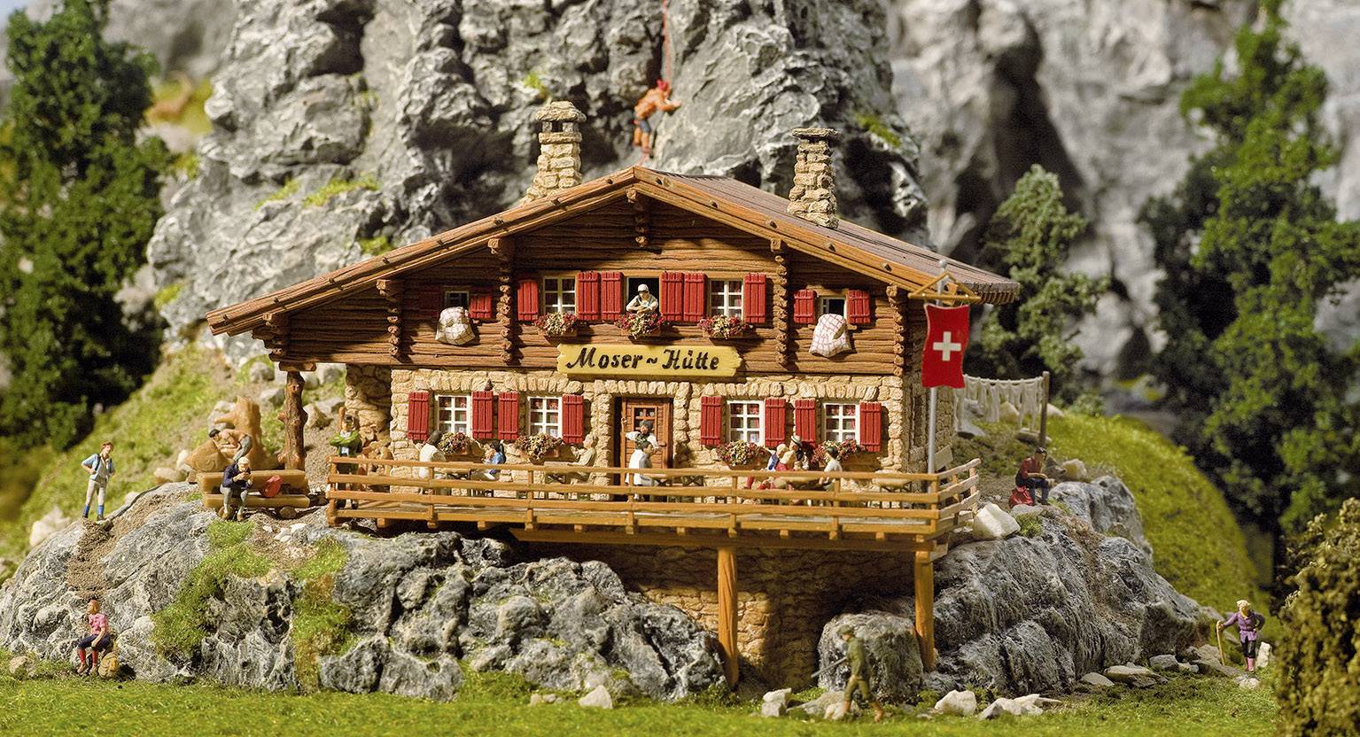 Et miniature-bjergchalet med påskriften 'Moser-Hütte', omgivet af klipper og træer, med et schweizisk flag.