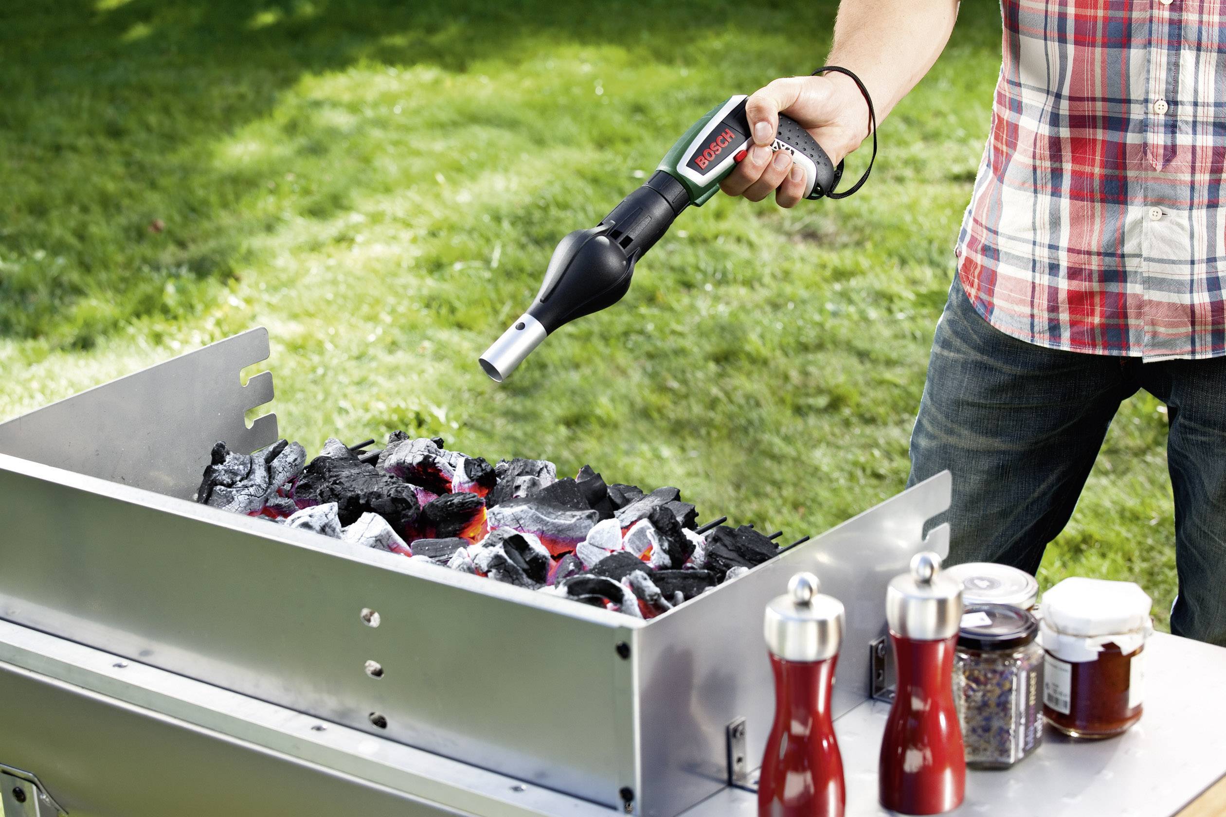 En person tænder grillkul med en elektrisk optændingsanordning. Ved siden af står krydderiflasker og glasbeholdere på et bord.