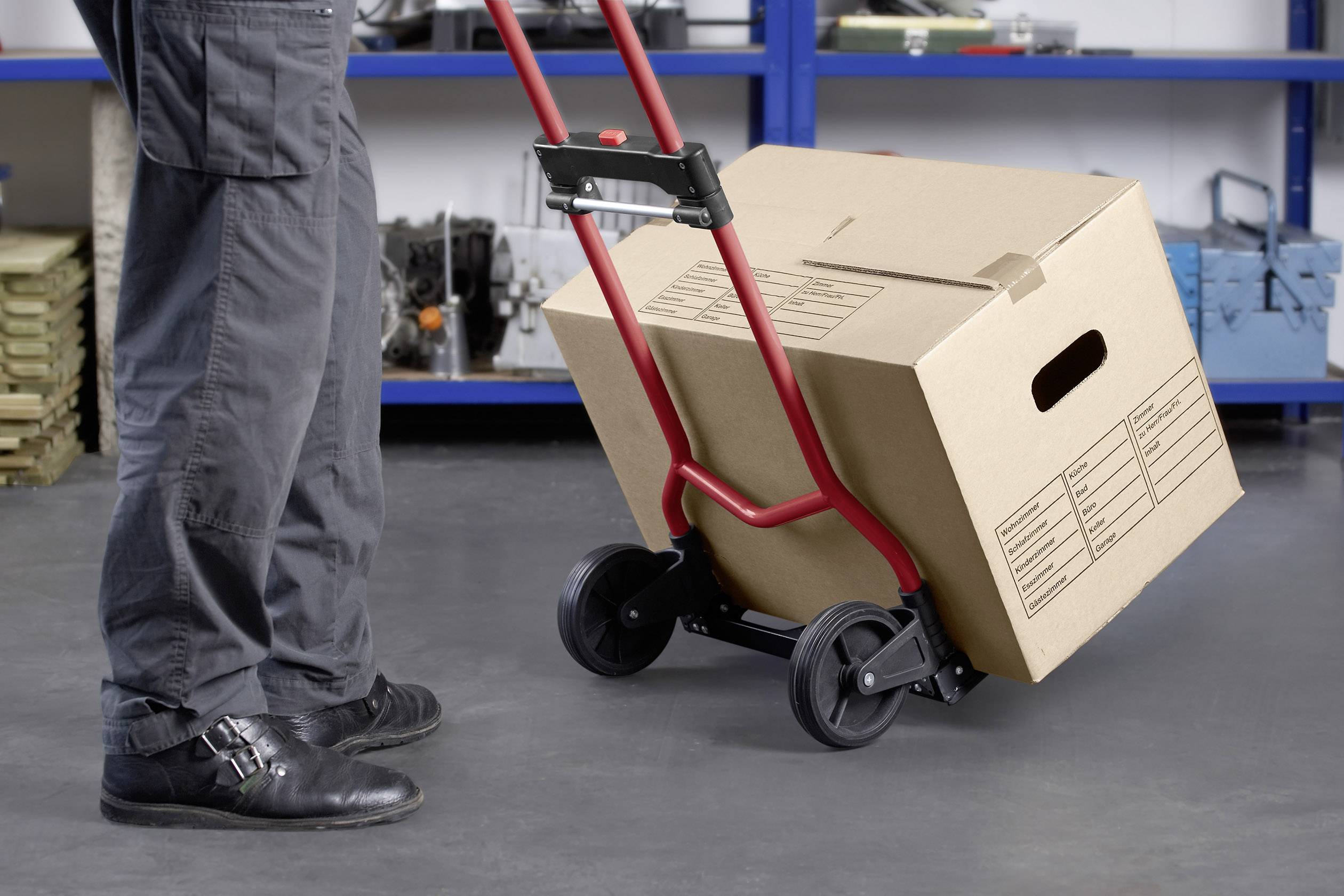 En person transporterer en stor papkasse på en håndvogn i et rum med blå hylder i baggrunden.