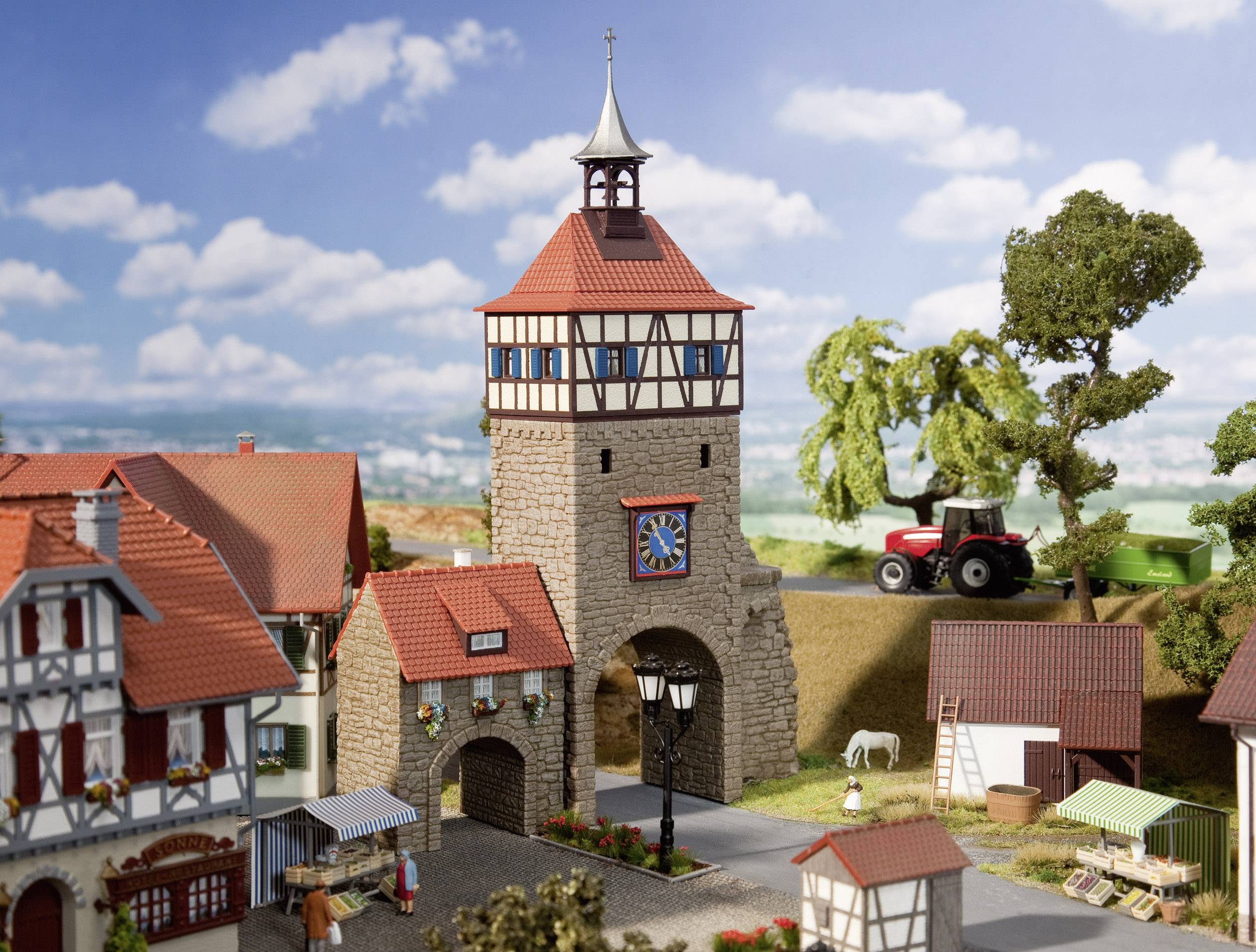 Lille miniaturelandsby med bindingsværkshuse og en port med et tårn; i baggrunden en rød traktor på en mark under en blå himmel.
