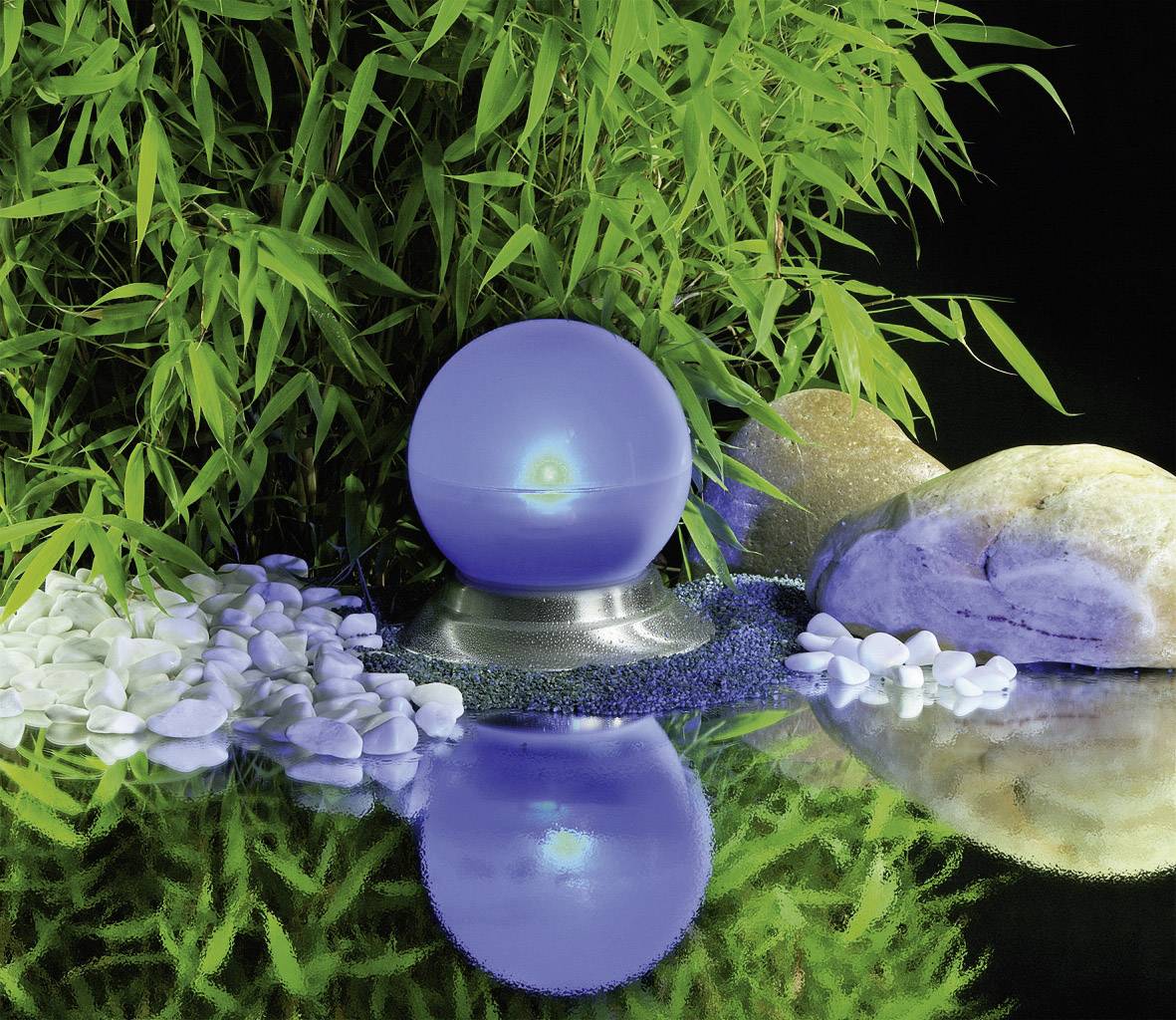 En blå glaskugle ligger på en piedestal, omgivet af grønne bambusblade, hvide kæselsten og reflekteres i vandet.