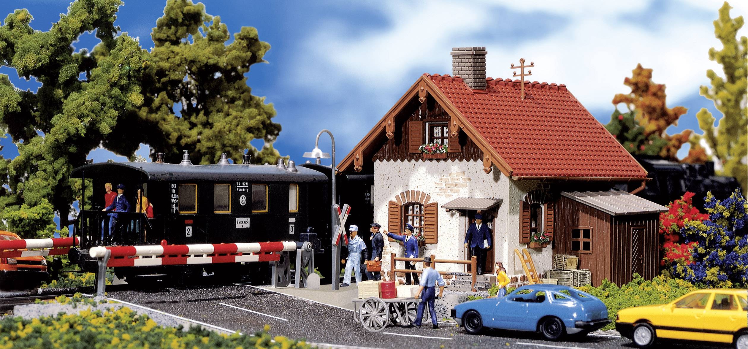 En miniature-jernbanescene med et sort tog ved en jernbaneoverskæring, omgivet af mennesker, et hus, biler og træer.
