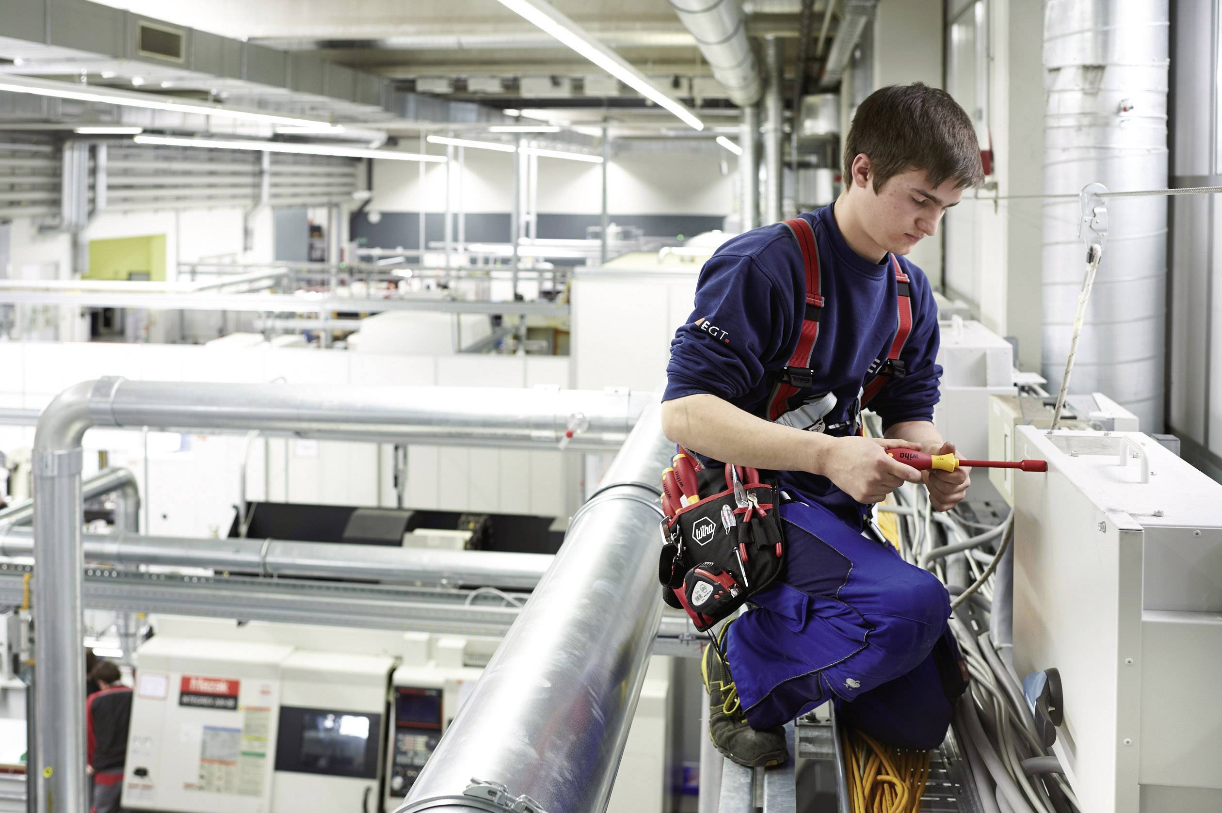 En person arbejder i en fabrikshal med elektriske apparater. Vedkommende bærer arbejdstøj og sikkerhedsudstyr.