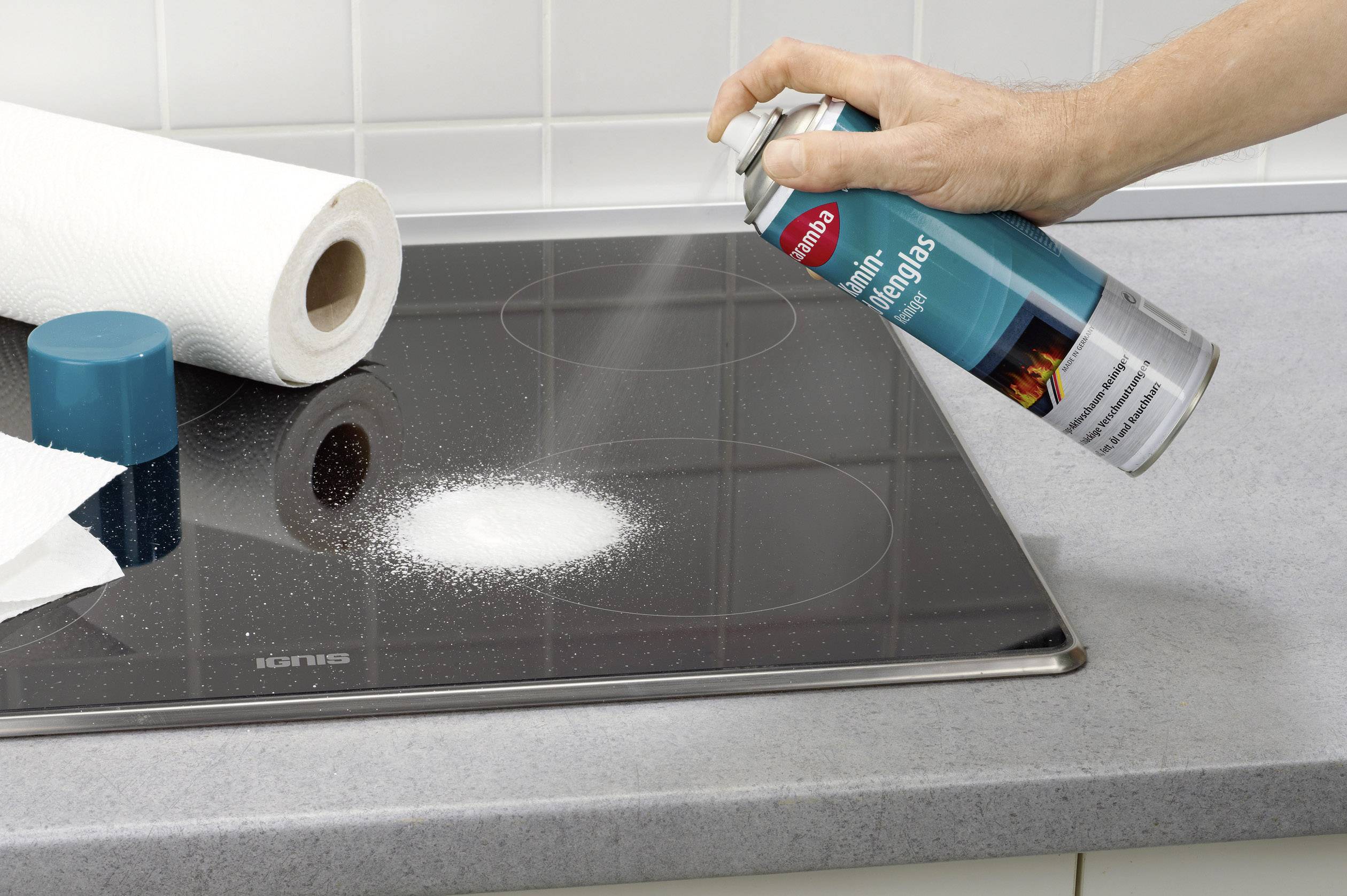 En person sprayer rengøringsmiddel på et snavset keramisk kogefelt. Ved siden af ligger køkkenrulle og en rengøringssvamp.