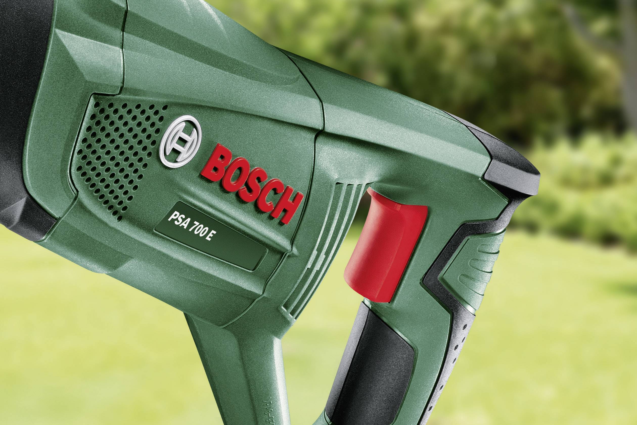 En grøn stiksav fra mærket Bosch, model PSA 700 E, med en rød kontakt. I baggrunden ses en sløret havelandskab.