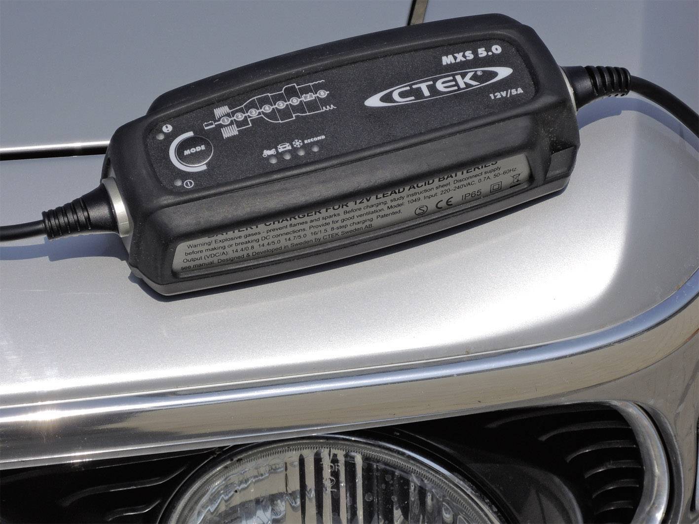 'CTEK MXS 5.0' autobatterilader ligger på en motorhjelm. Enheden viser forskellige opladningscyklusser og driftslamper.