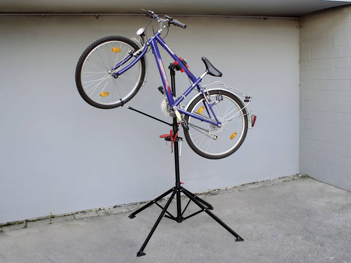En cykel står lodret på en sort reparationsstander i et betonrum. Standeren holder cyklen fast ved bagakslen.