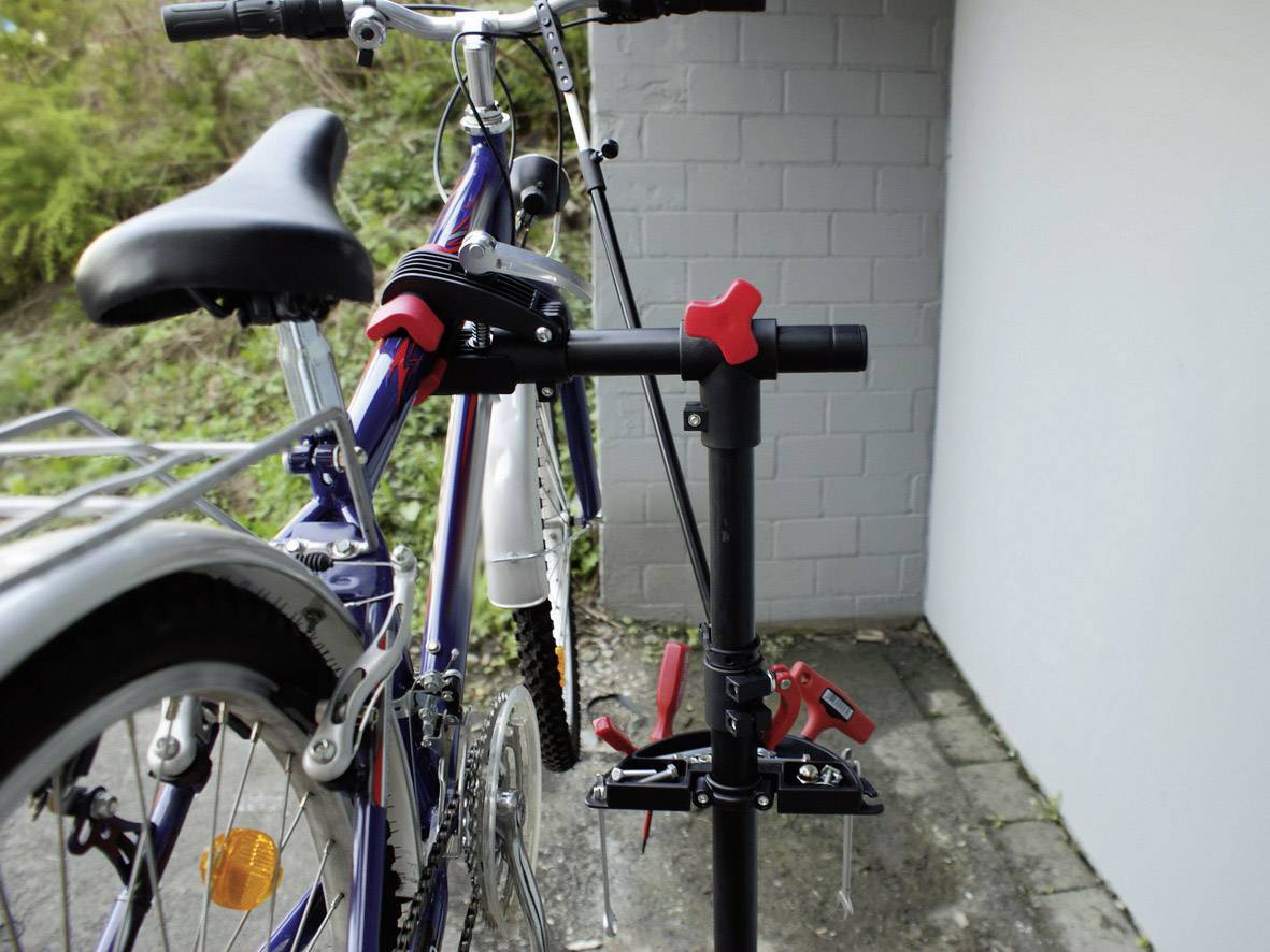 En cykel står på et stativ foran en hvid væg, klar til vedligeholdelse. Baghjulet er løftet op, røde beslag er synlige.