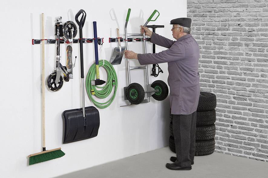 En ældre mand arrangerer værktøj og haveredskaber på en garagevæg. Til venstre hænger en kost og en sneskraber, til højre læner dæk sig op ad væggen.
