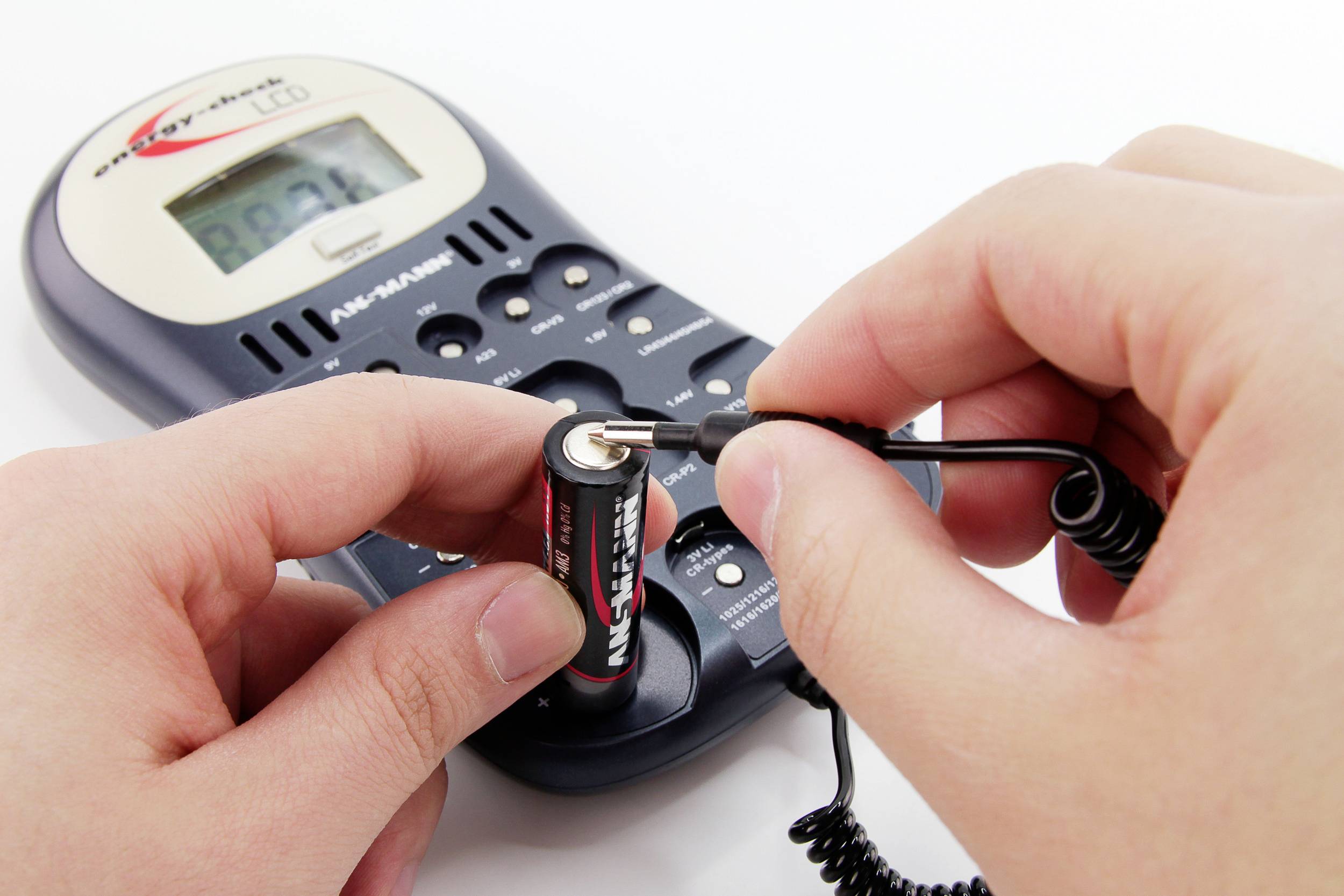 Eine Person hält eine AA-Batterie und misst die Spannung mit einem Messgerät. Im Hintergrund ist ein Ladegerät zu sehen.
