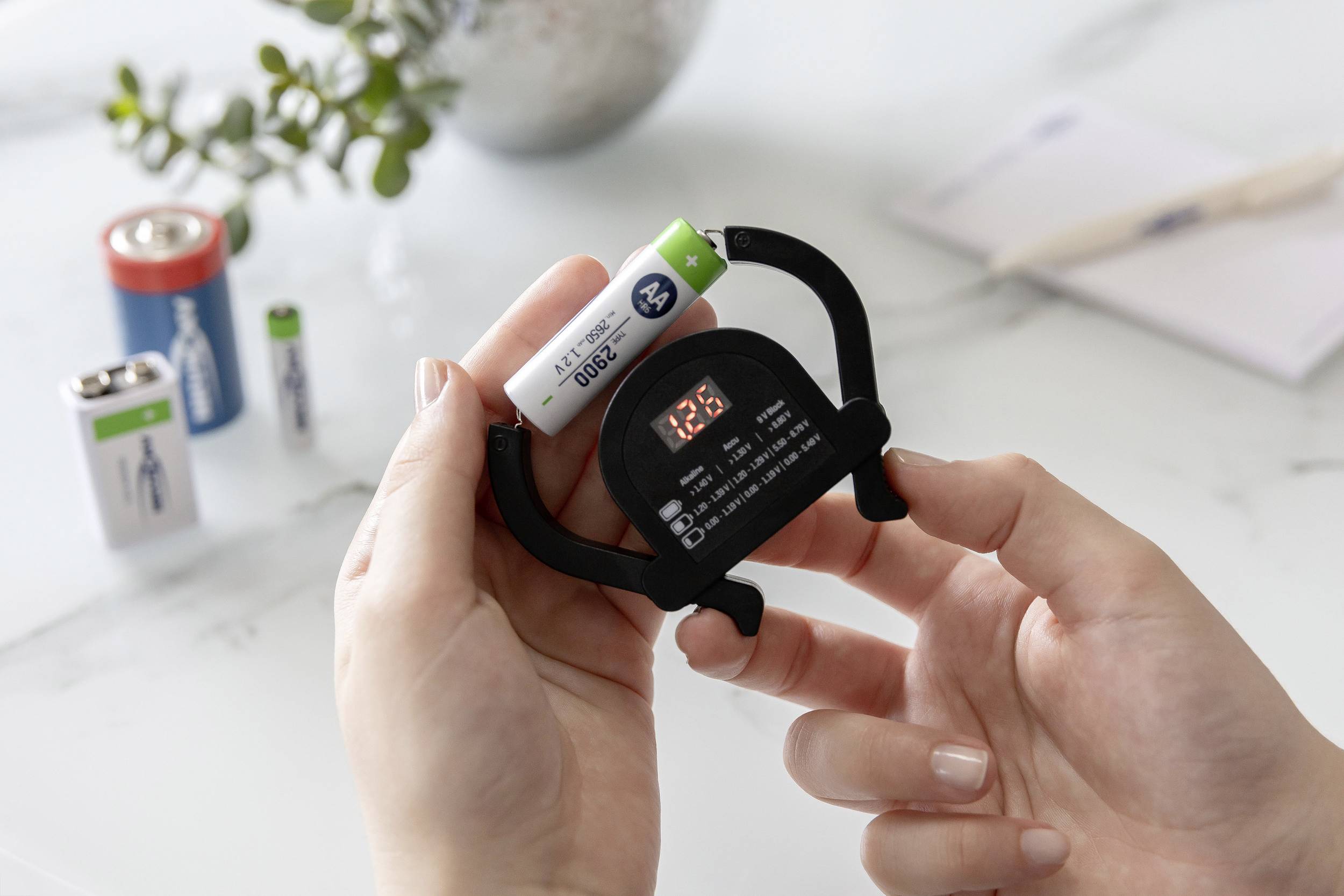 Eine Person misst die Spannung einer AA-Batterie mit einem digitalen Spannungsmesser. Weitere Batterien liegen im Hintergrund.