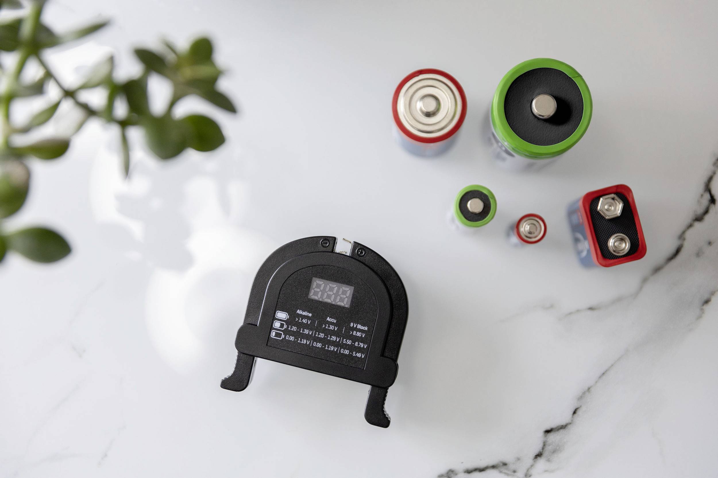 Ein schwarzes Batteriemessgerät liegt auf einem Tisch, umgeben von verschiedenen Batterien. Links sind verschwommene Blätter sichtbar.