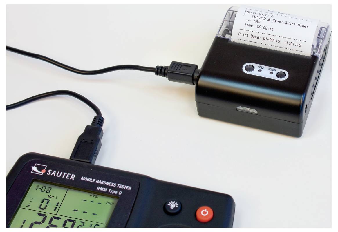 'Digitales Härteprüfgerät mit Drucker, zeigt Messergebnisse an, verbunden durch USB-Kabel. Bildschirm und Drucker im Vordergrund.'