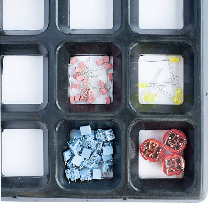 Organizerfach mit elektronischen Komponenten: rosa Kondensatoren, gelbe LED-Leuchten, blaue Kondensatoren und zwei rote Leiterplatten.