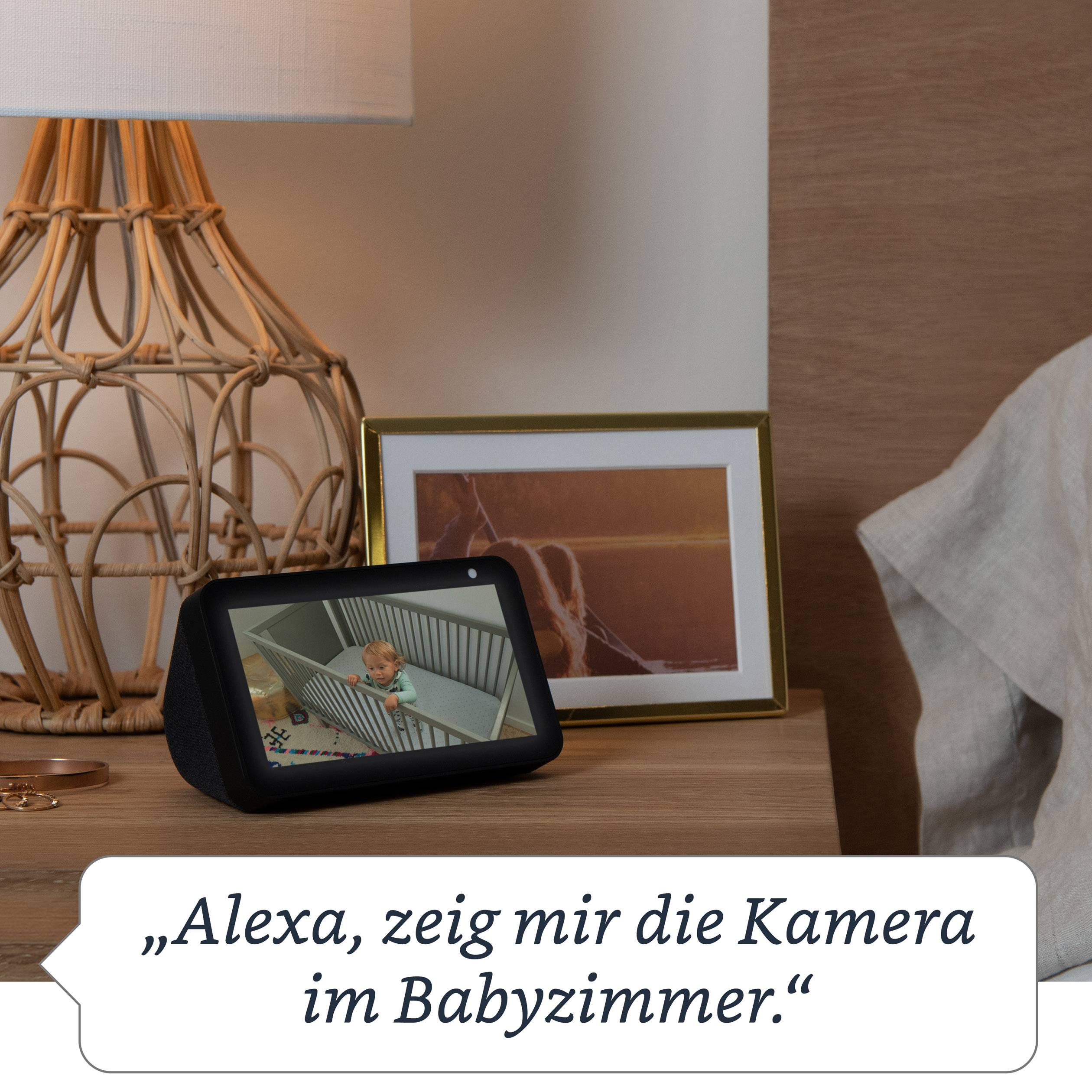 Ein sprachgesteuertes Gerät auf einem Nachttisch zeigt ein Baby im Laufstall. Darunter steht: 'Alexa, zeig mir die Kamera im Babyzimmer.'