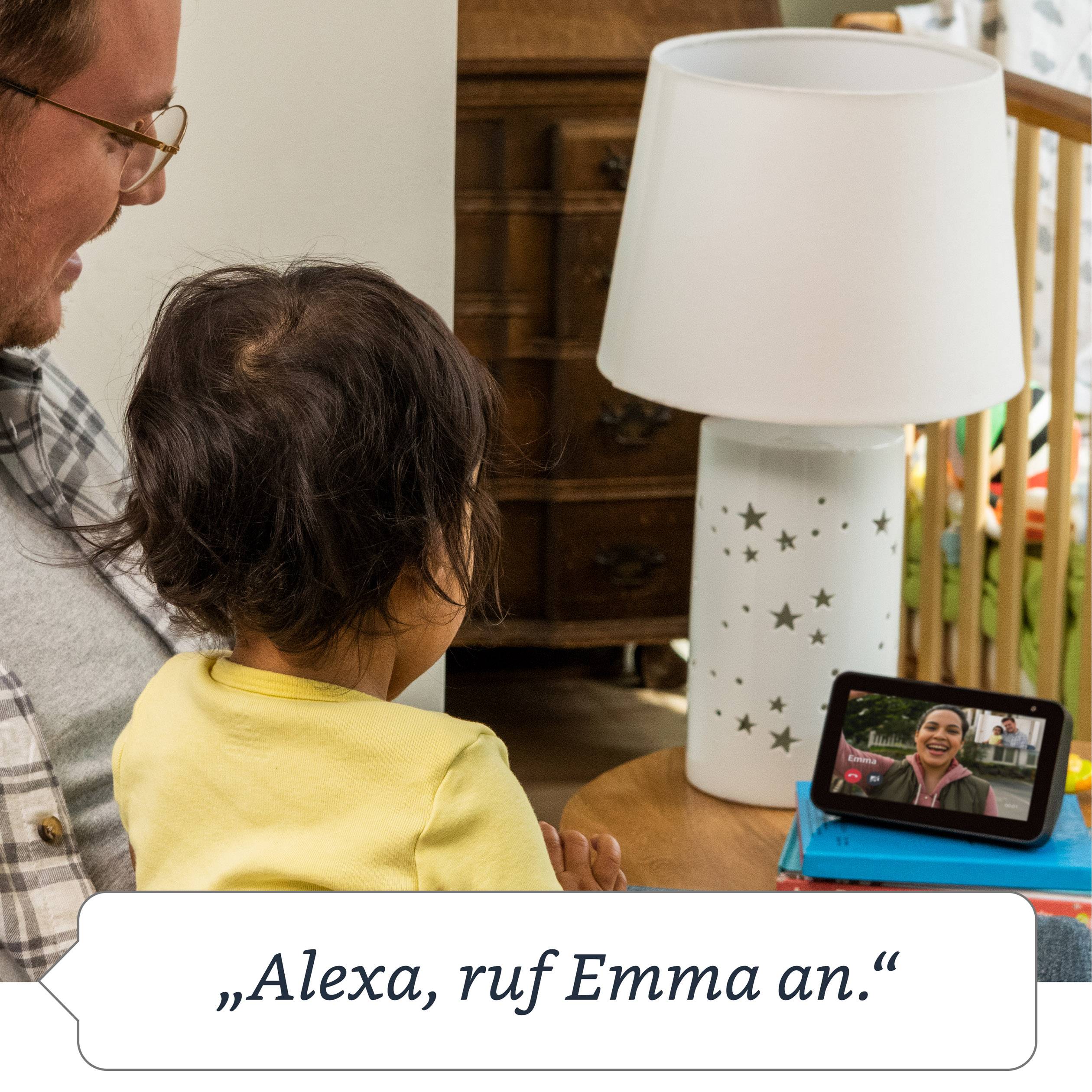 Ein Mann mit einem Kind sitzt neben einem Tisch mit einem Tablet, auf dem ein Videoanruf läuft. Der Text lautet: „Alexa, ruf Emma an.“