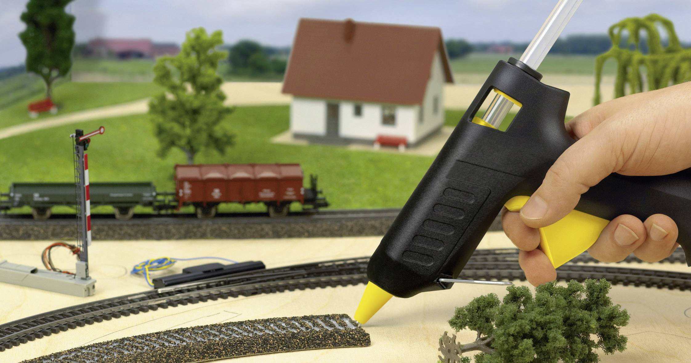 Eine Hand trägt mit einer Heißklebepistole Klebstoff auf eine Modelleisenbahnlandschaft auf, einschließlich Gleisen, Bäumen, Zugwagen und Gebäuden im Hintergrund.