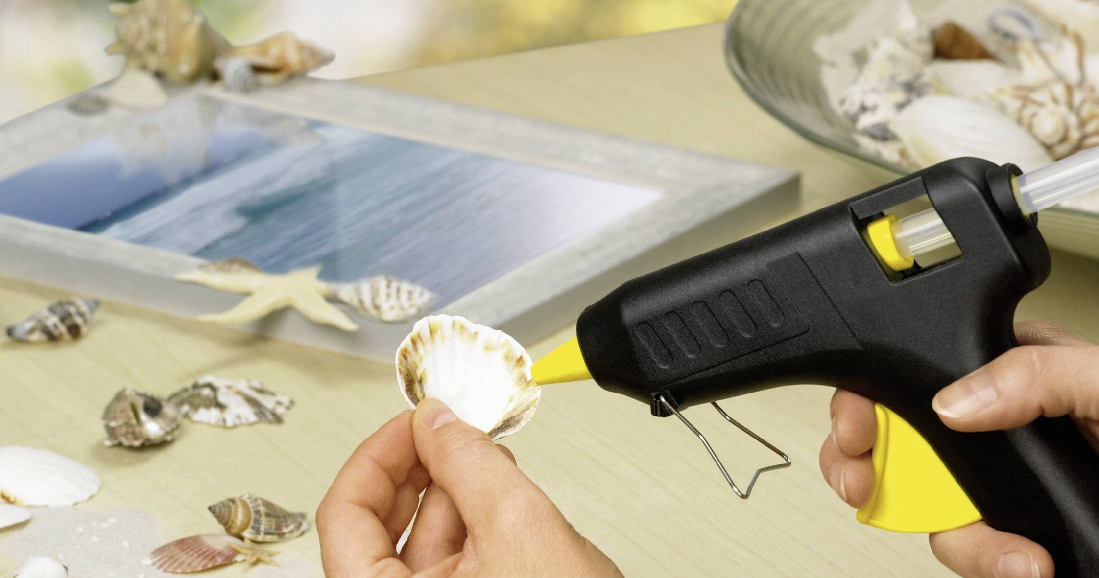 Eine Person verwendet eine Heißklebepistole, um eine Muschel an einen Bilderrahmen zu befestigen, der mit anderen Muscheln und einem Seestern auf einem Tisch dekoriert ist.