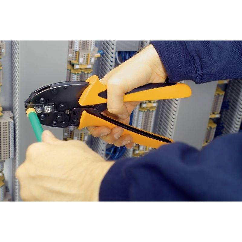 Eine Person verwendet eine orange-schwarze Crimpzange, um ein Kabel in einem Schaltschrank zu bearbeiten.