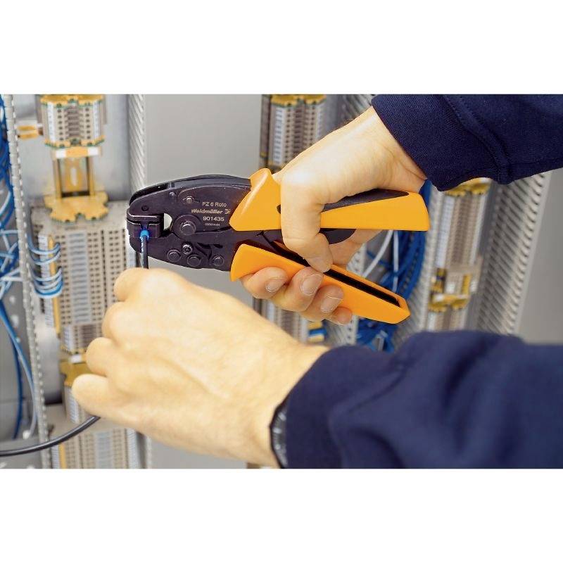 Eine Person crimpt ein Kabel mit einer orangenen Crimpzange in einem Schaltschrank voller Kabel und Anschlüsse.