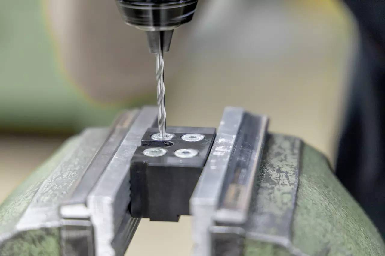 Ein Bohrer erstellt Löcher in einem Metallblock, der in einer Schraubzwinge befestigt ist und dabei präzises Bearbeiten in einem industriellen Umfeld betont.