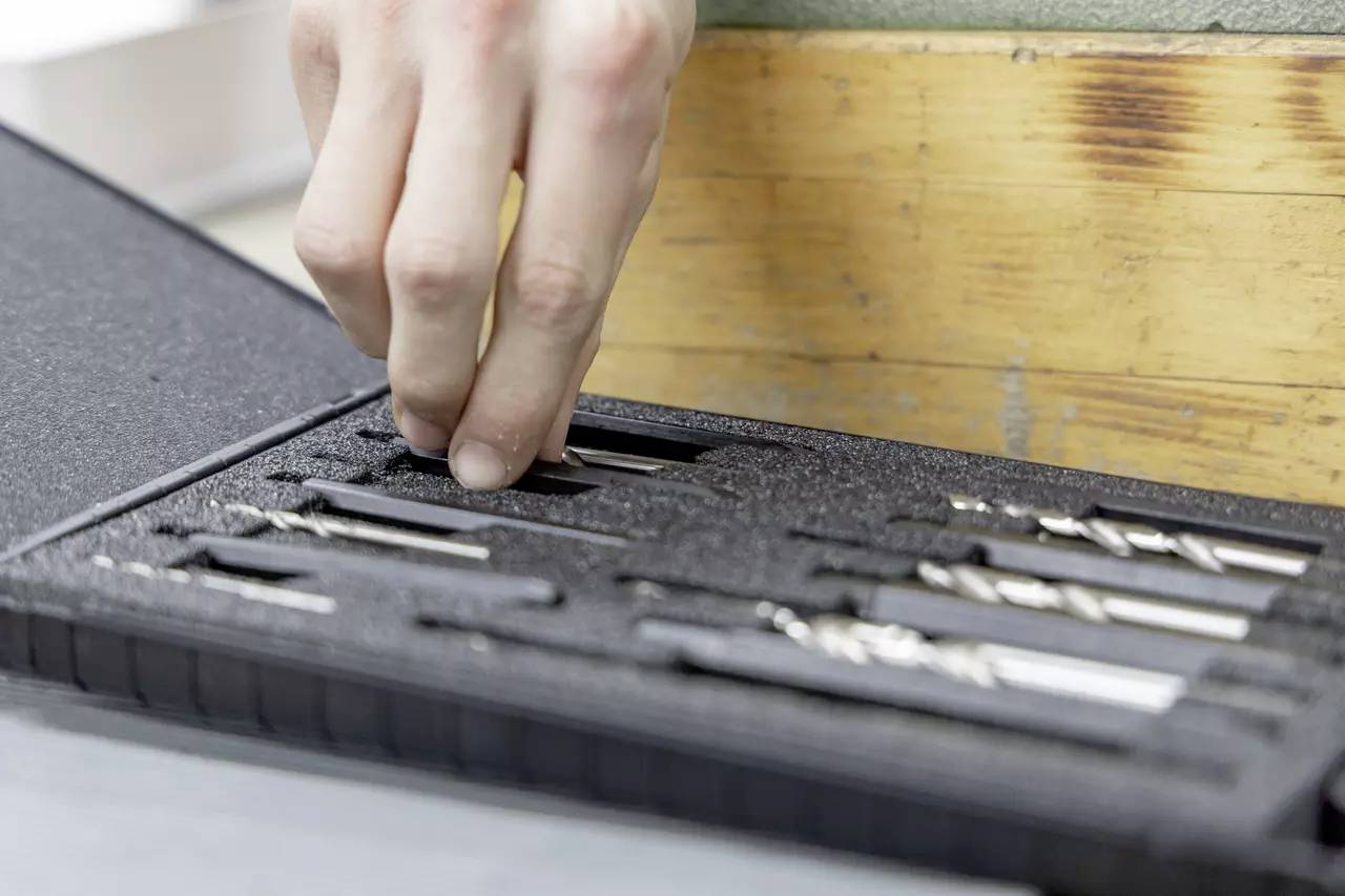 Eine Hand nimmt einen Bohrer aus einem mit Schaumstoff ausgelegten Etui, das verschiedene Metallbohrer enthält und auf einer Werkbank liegt.